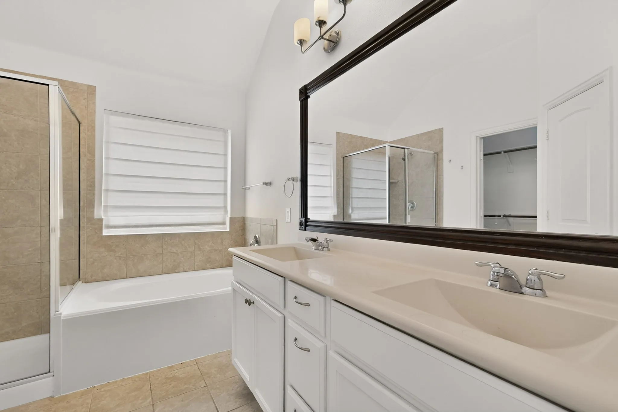 Dual-sink vanity with white cabinetry and a solid-surface countertop, oversized framed wall mirror, soaking tub, glass-enclosed shower, and tile flooring