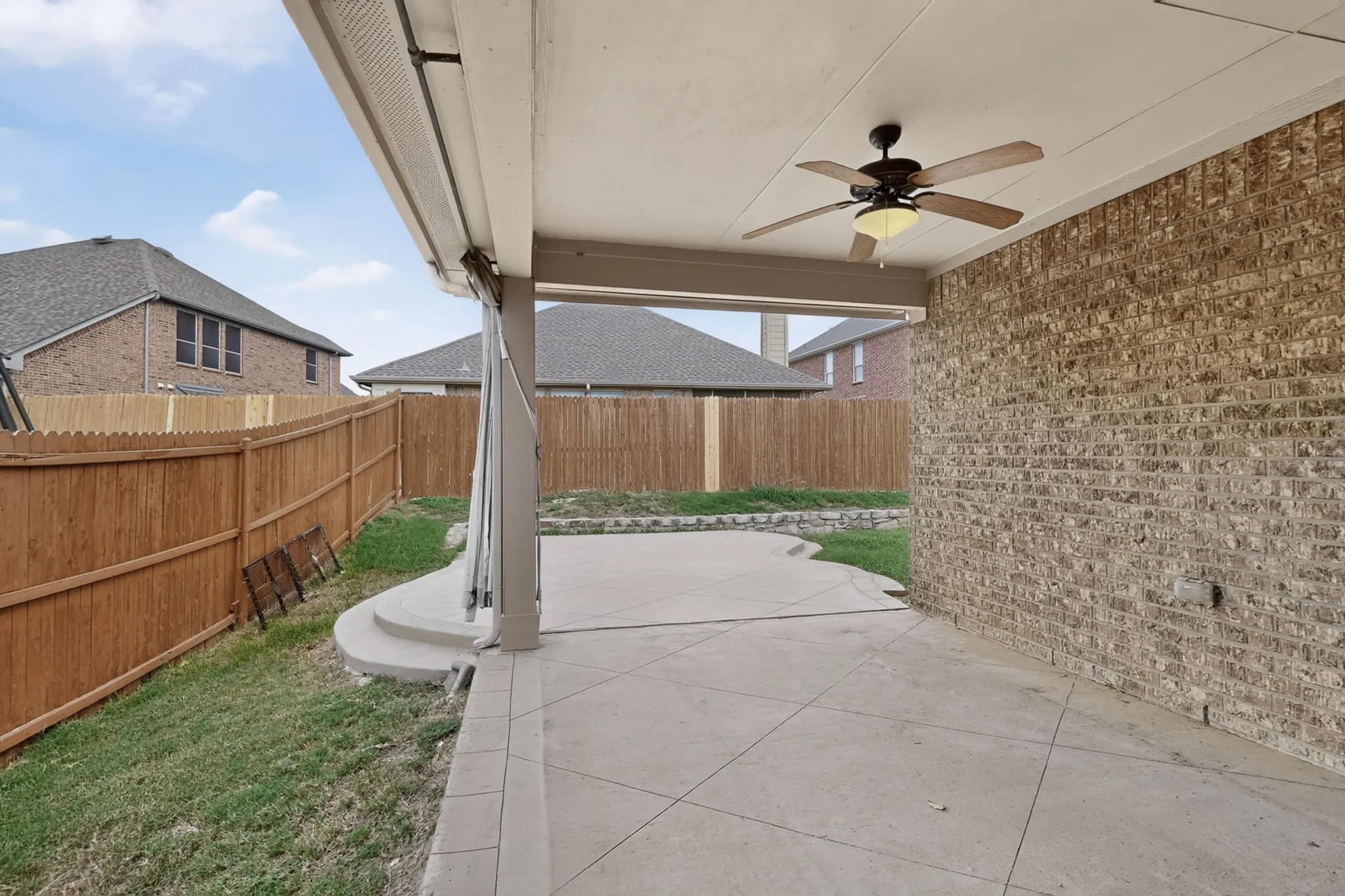 Covered patio featuring scored concrete flooring, a ceiling fan, a brick exterior wall, and a wood privacy fence
