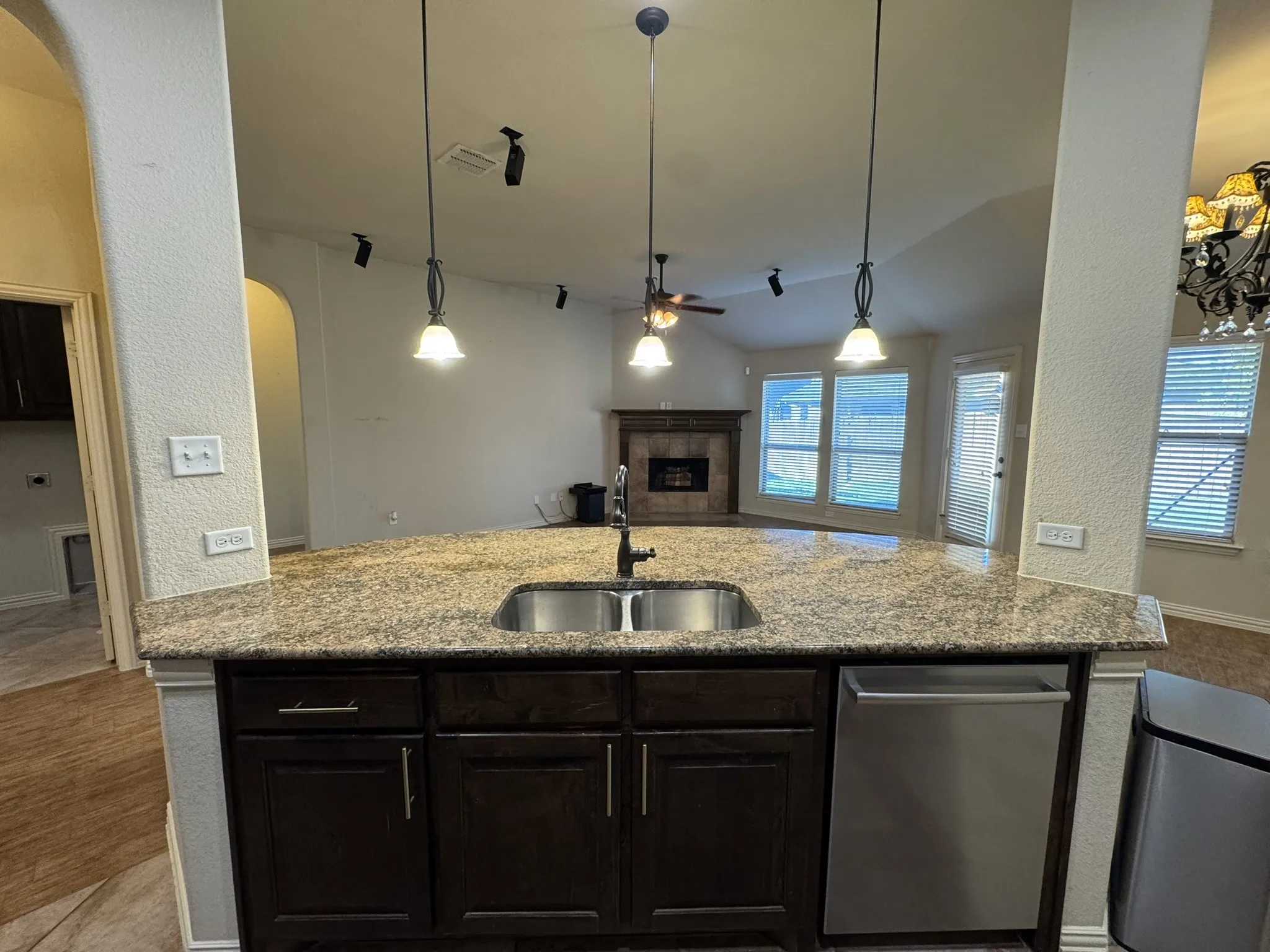 Kitchen island featuring stone-surfaced countertops, stainless steel undermount double-basin sink, dark wood cabinetry, three pendant light fixtures, and a stainless steel dishwasher