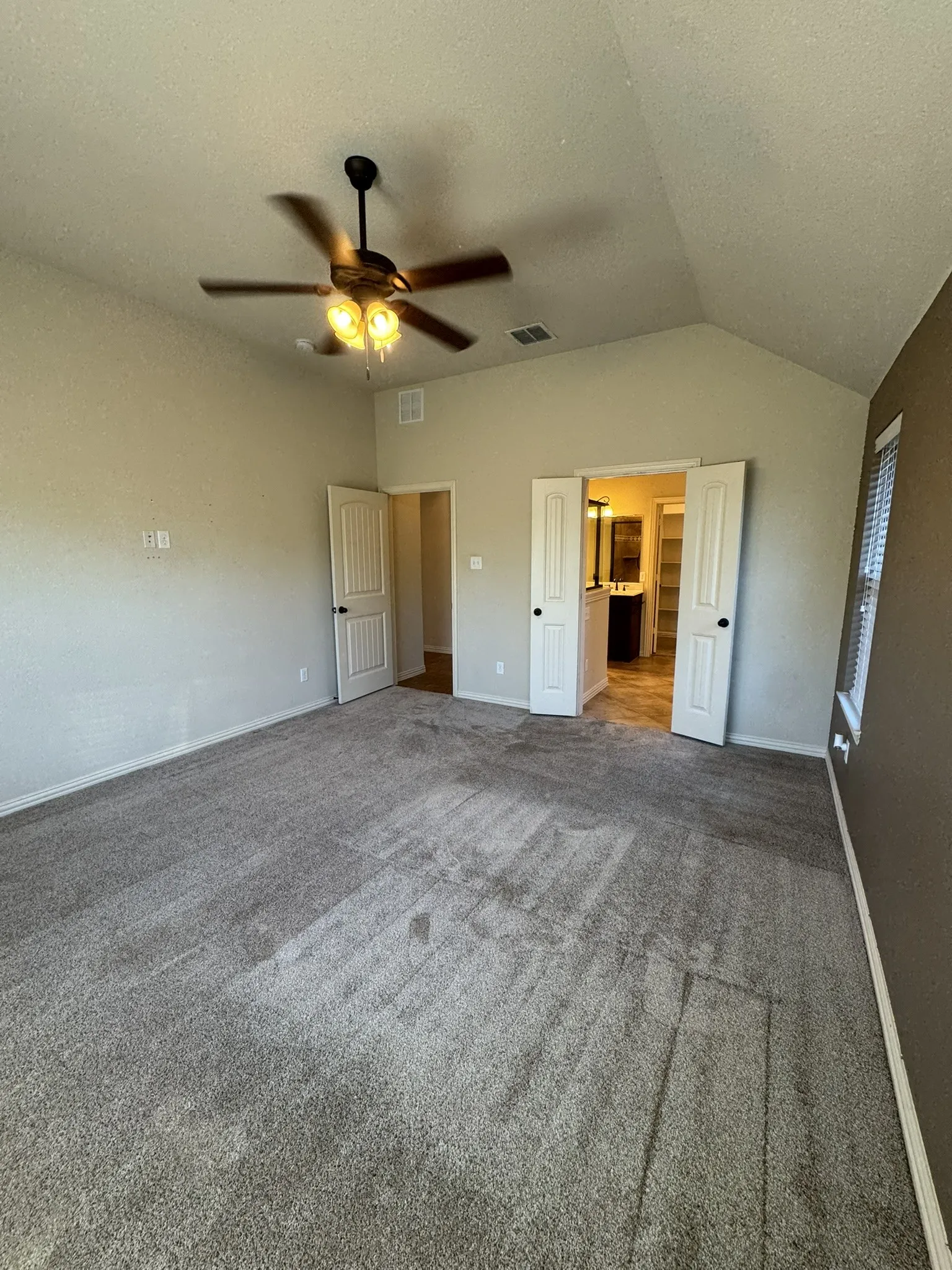 Vaulted ceilings featuring a central ceiling fan with integrated lighting, neutral wall tones, carpeted flooring, and double doors opening to an auxiliary space