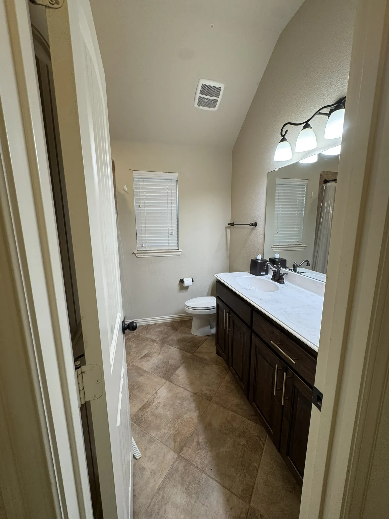 Dark wood-finish cabinetry featuring dual-basin vanity with a solid surface countertop, ceramic tile flooring, and vaulted ceiling
