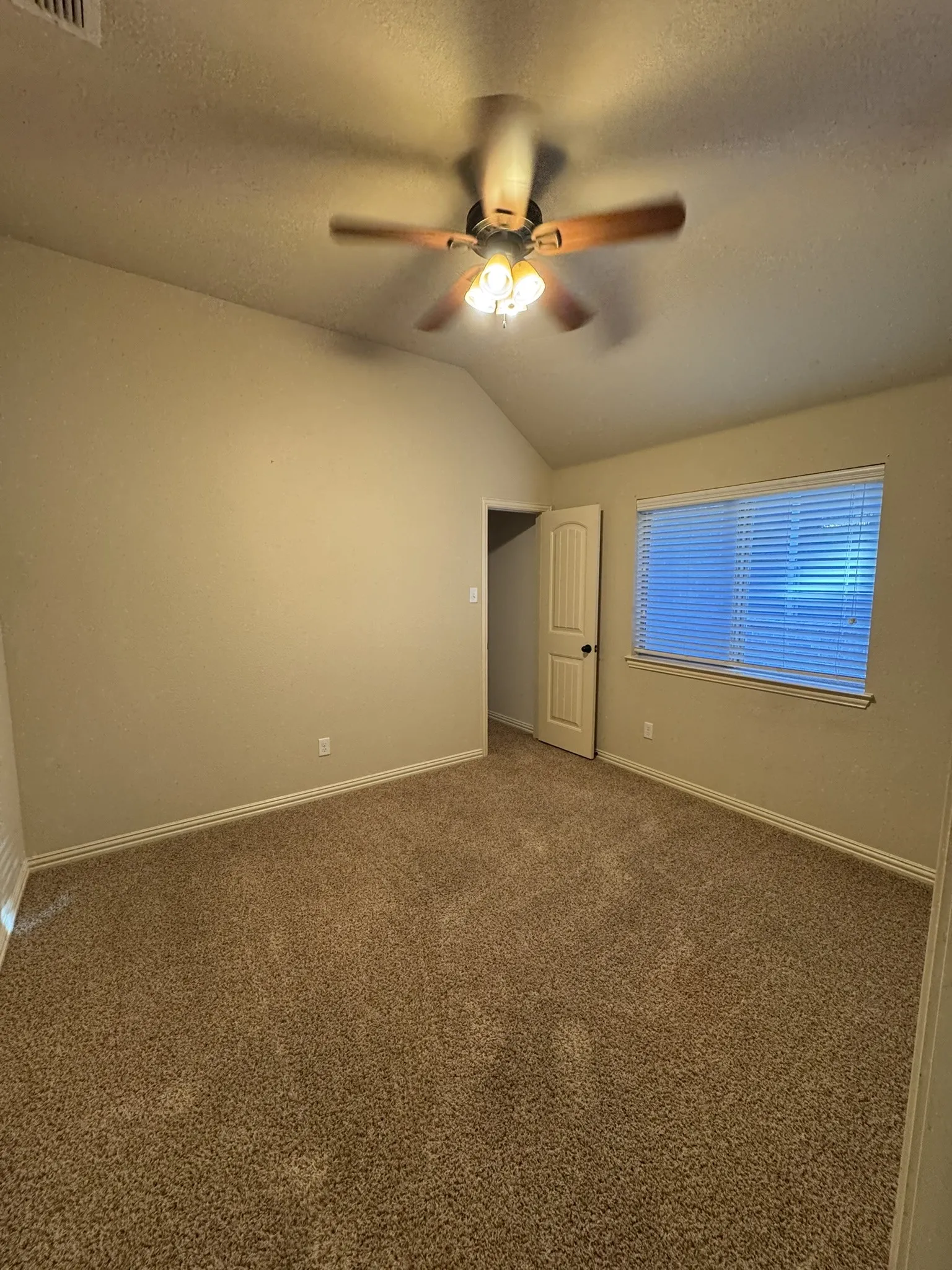 Vaulted ceiling with a centrally mounted ceiling fan featuring integrated tri-bulb lighting, neutral carpeting, and window with horizontal blinds