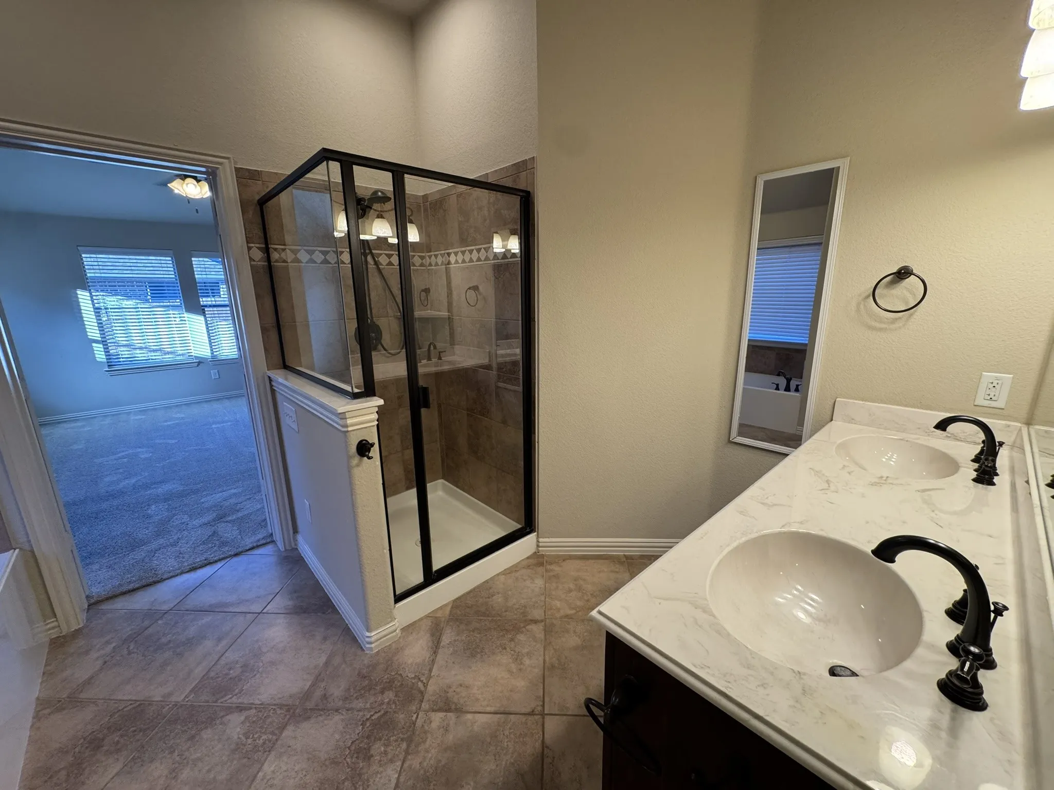 Bathroom featuring dual-sink vanity with stone-finish countertop, dark metal fixtures, tiled flooring, and a walk-in shower with a black-frame enclosure