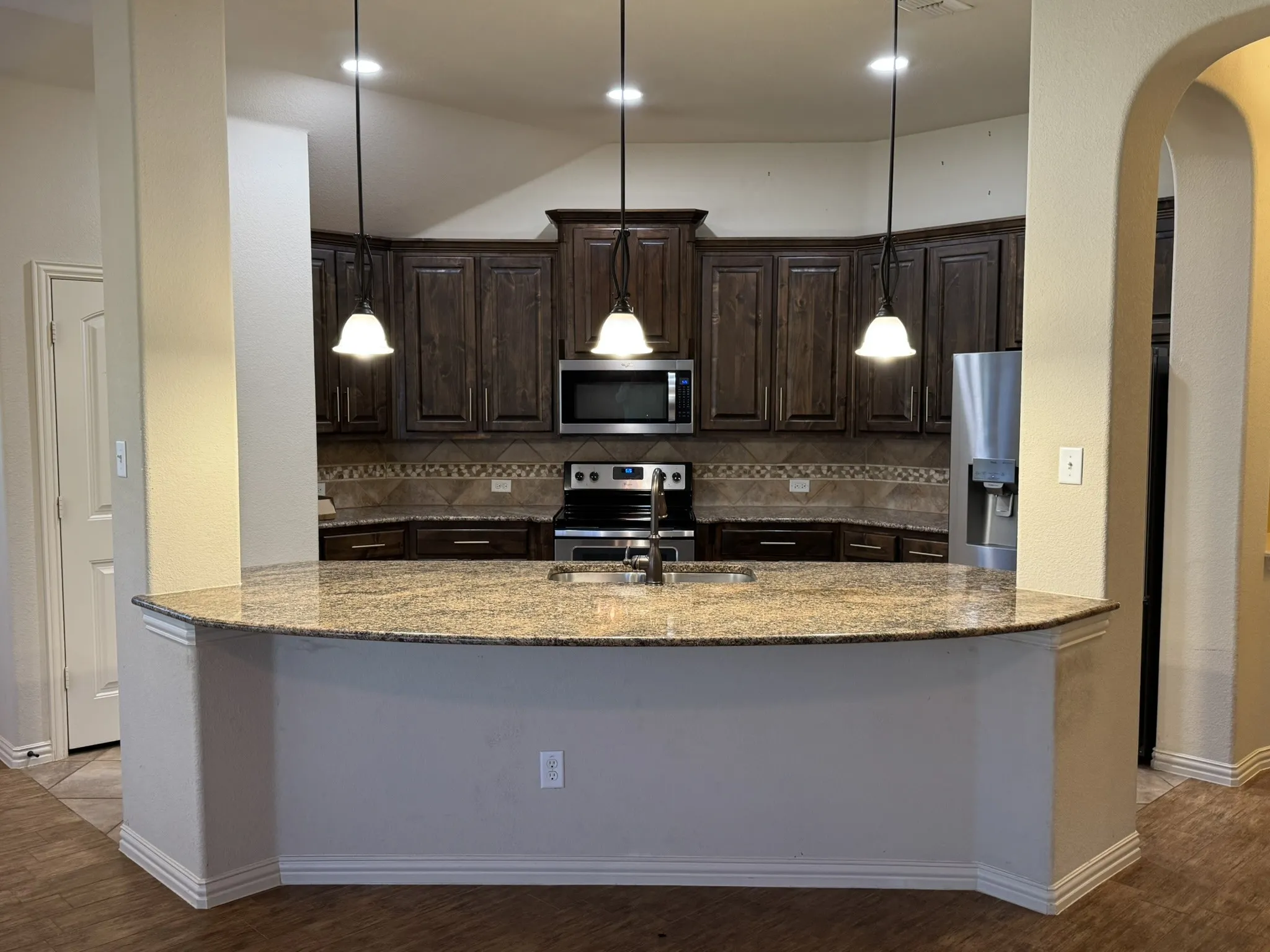 Kitchen featuring a curved stone-surface island, dark-toned wood cabinetry, built-in stainless steel appliances, decorative tile backsplash, and three pendant lights