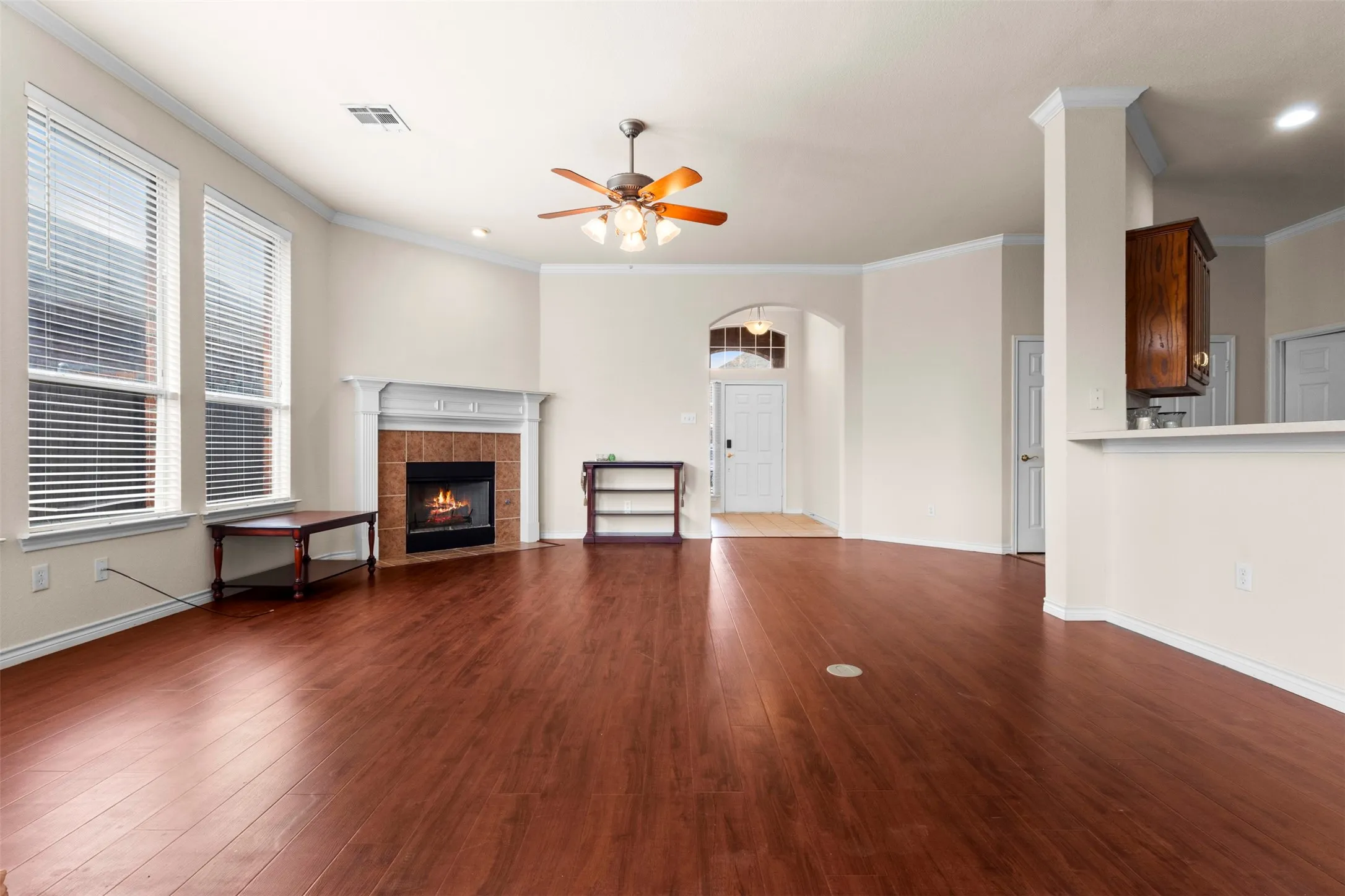 Wood-finish flooring, fireplace with a tile surround and mantel, arched entryway, crown molding, and a ceiling fan with integrated lighting