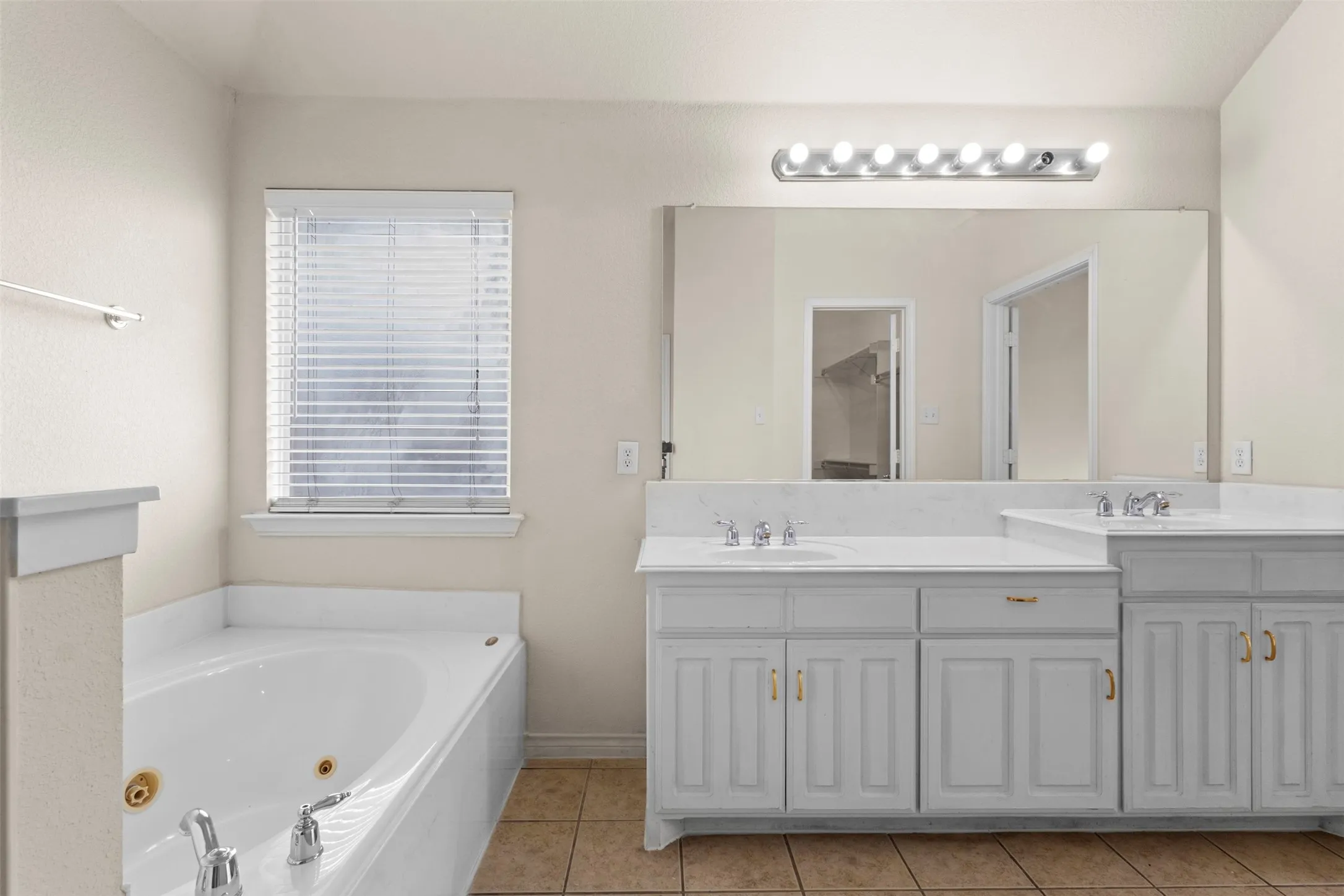 Dual-sink vanity with white cabinetry and stone-finish countertop, jetted soaking tub, oversized wall-mounted mirror, and tile flooring