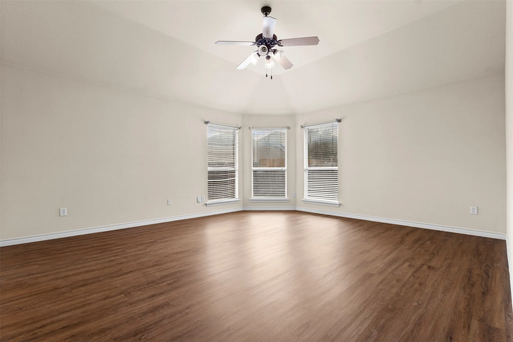 Vaulted ceiling featuring a central multi-blade ceiling fan, wood-finish flooring, and a bay of three windows