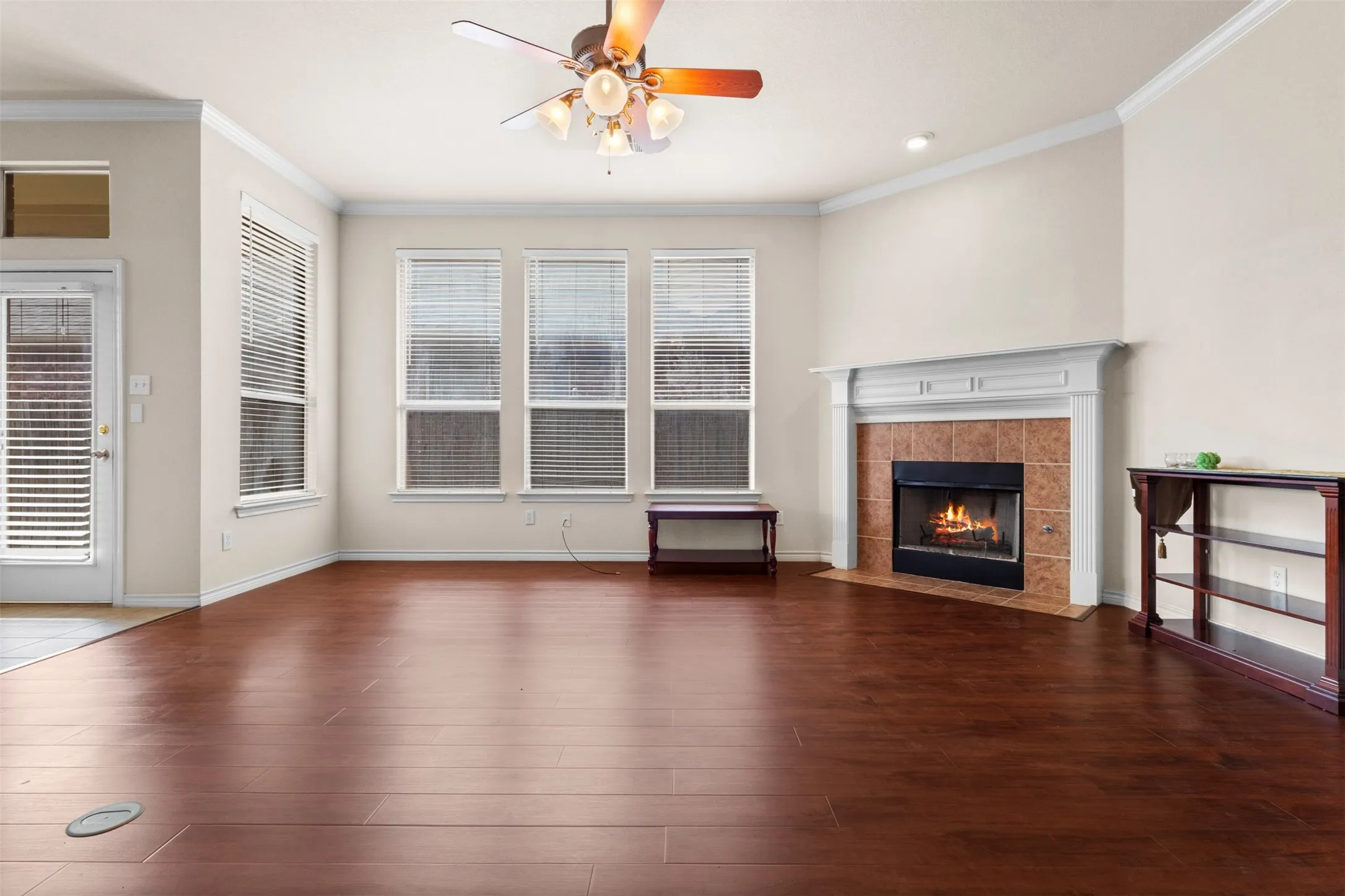Living area featuring wood-finish flooring, a fireplace with a white mantel and tile surround, crown molding, and multiple windows with horizontal blinds