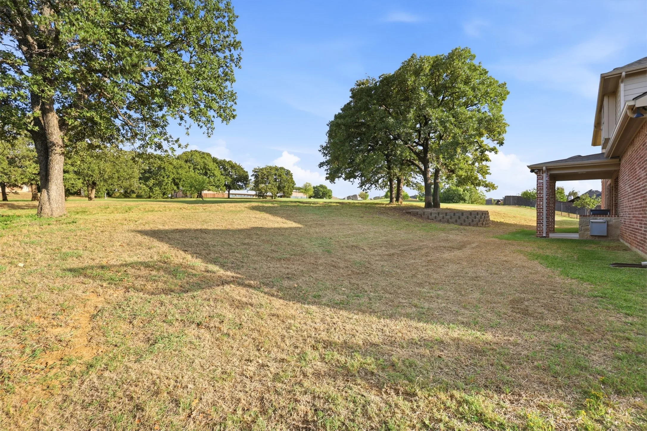 Expansive grounds featuring mature shade trees, a segmented retaining wall, and a covered brick patio