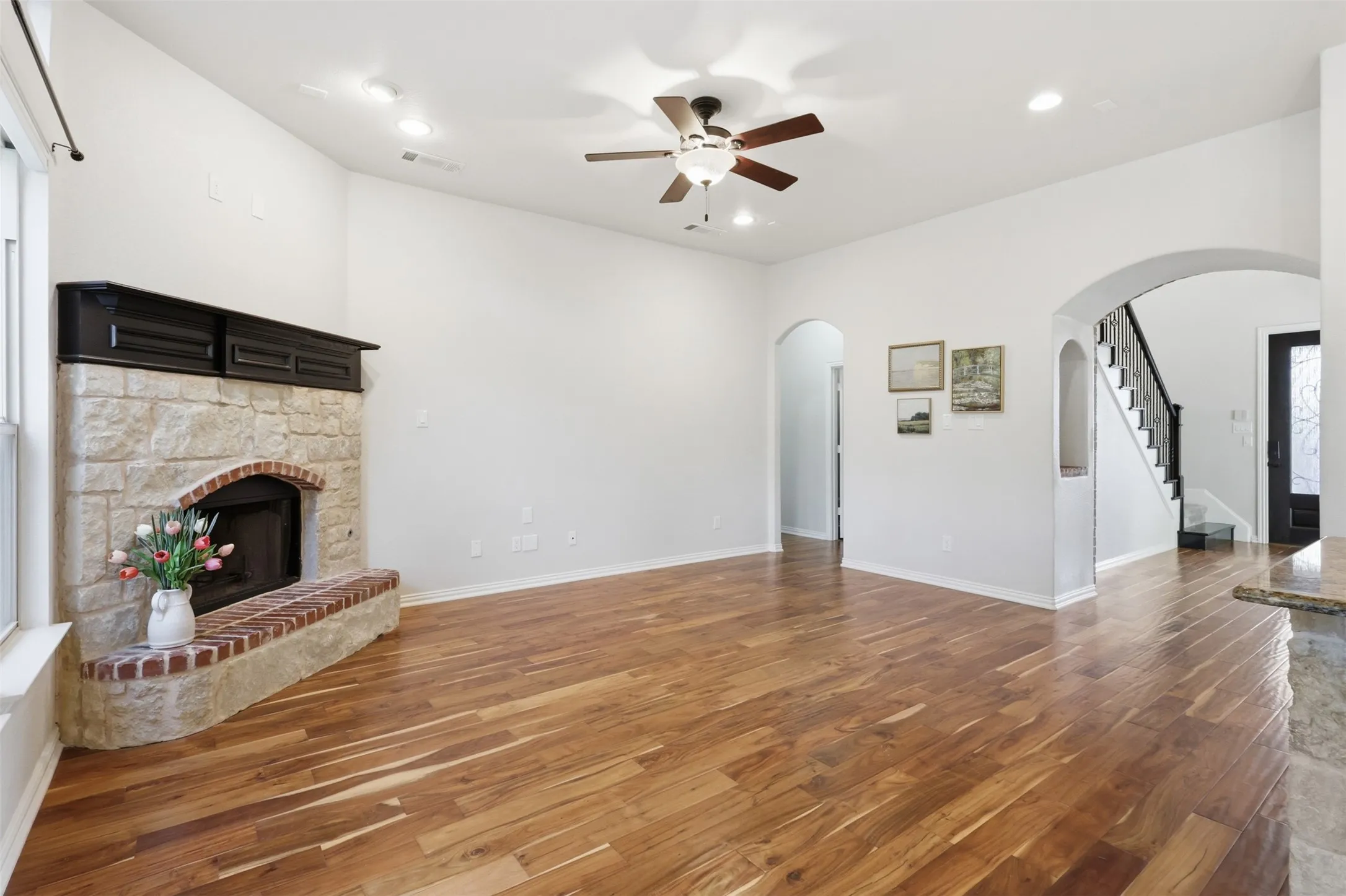 Living area featuring wood-finish flooring, a stone fireplace with a brick hearth, a ceiling fan, and arched architectural entryways