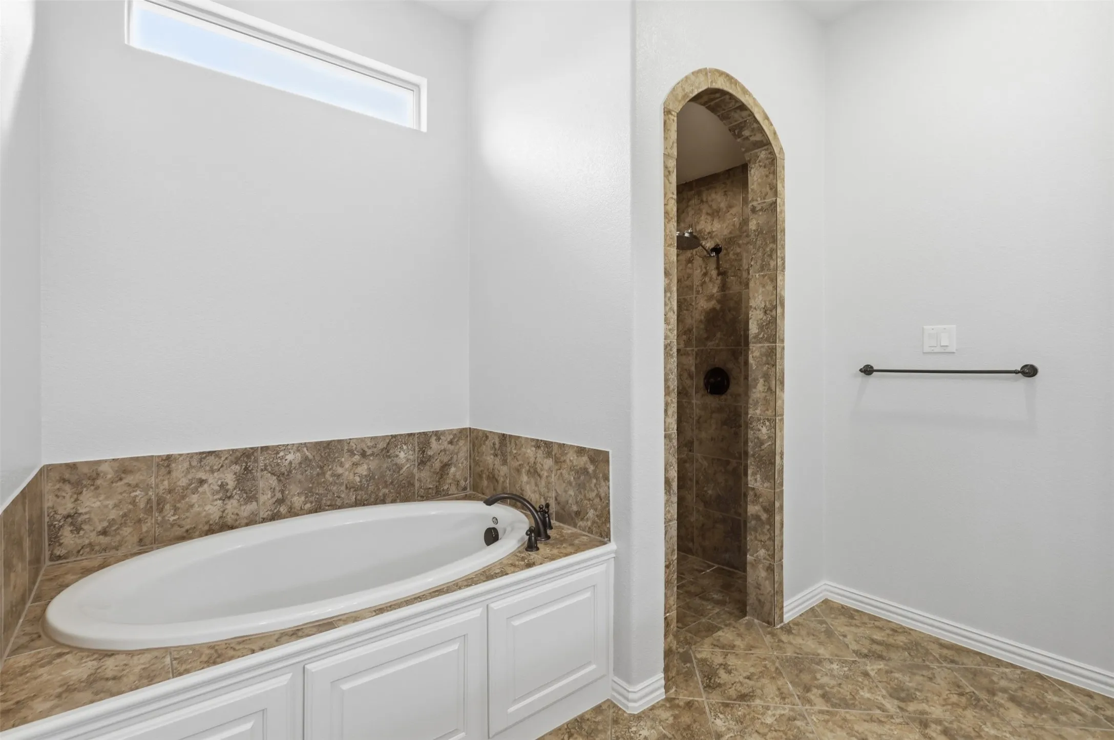Oval soaking tub with stone-finish surround, arched shower entry featuring tiled interior, transom window, and white cabinetry