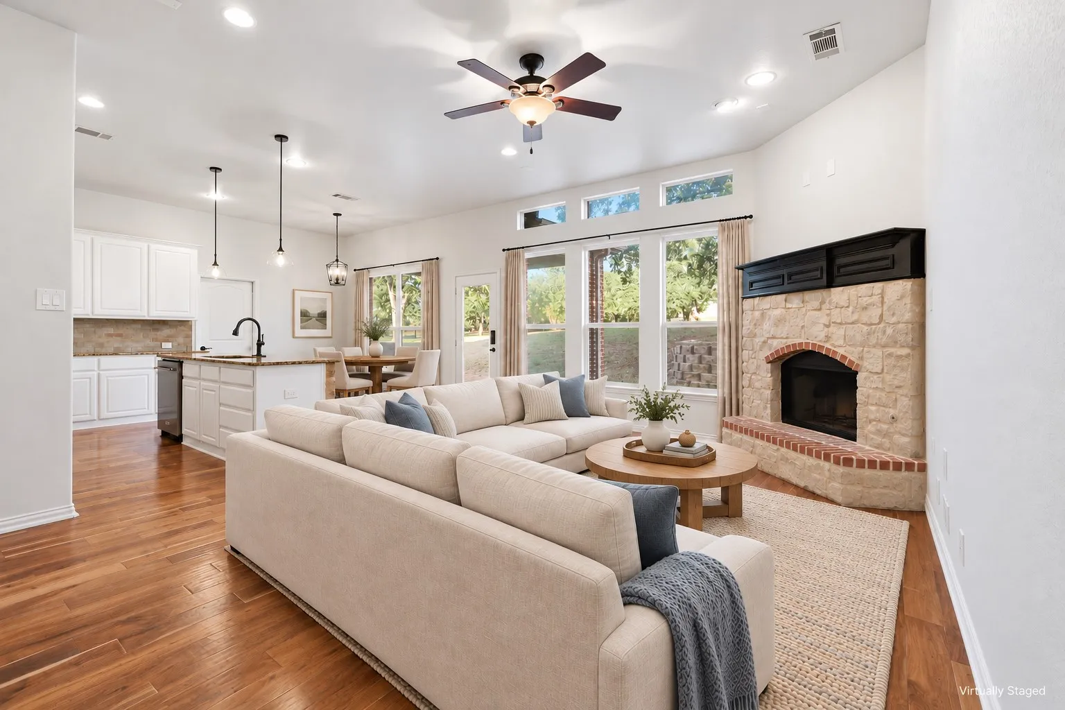 Open floor plan featuring wood-finish flooring, white shaker-style cabinetry, an island with pendant lighting, and a stone fireplace with a brick hearth