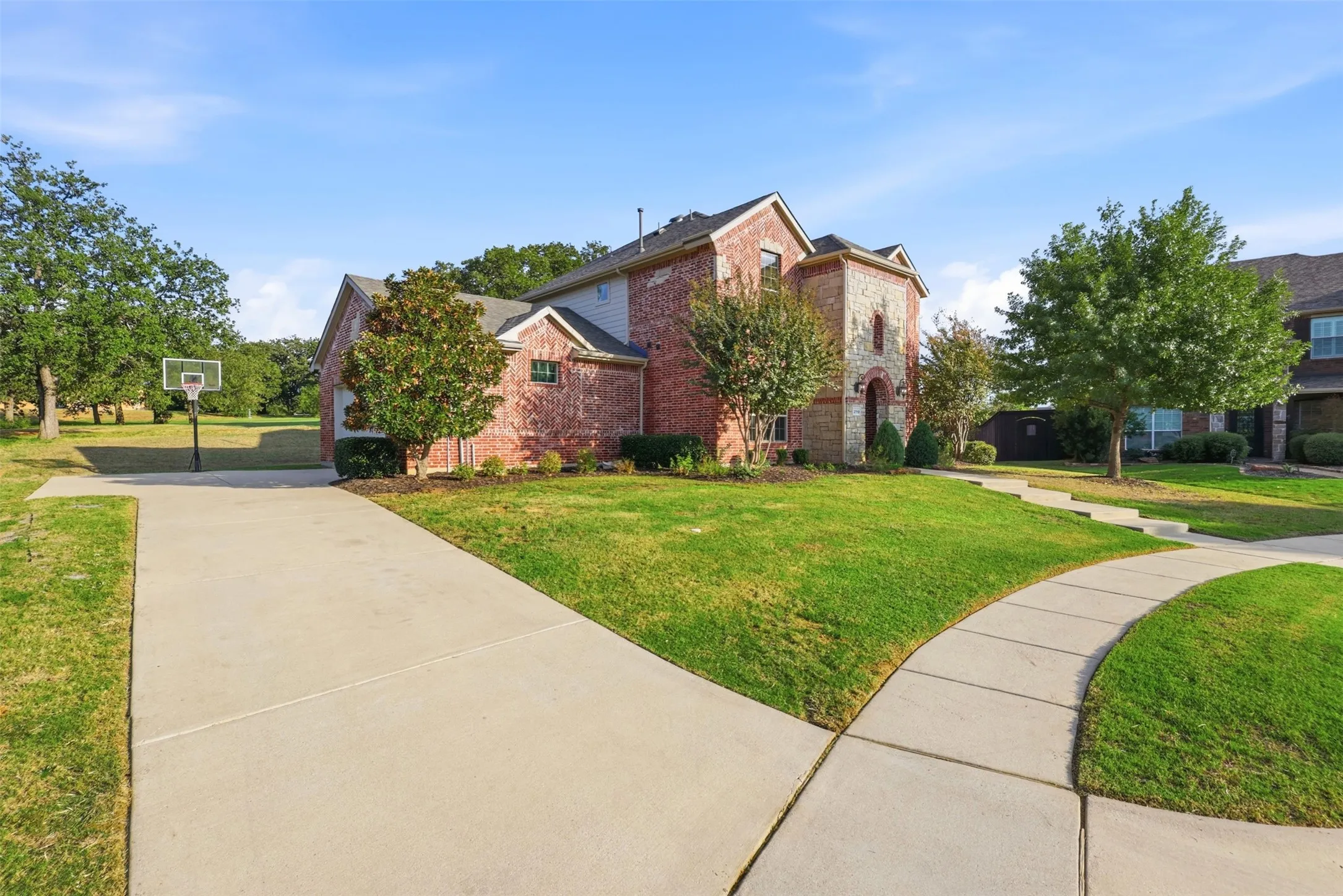 Two-story exterior featuring multi-toned brick and stone masonry, concrete driveway, and established landscaping