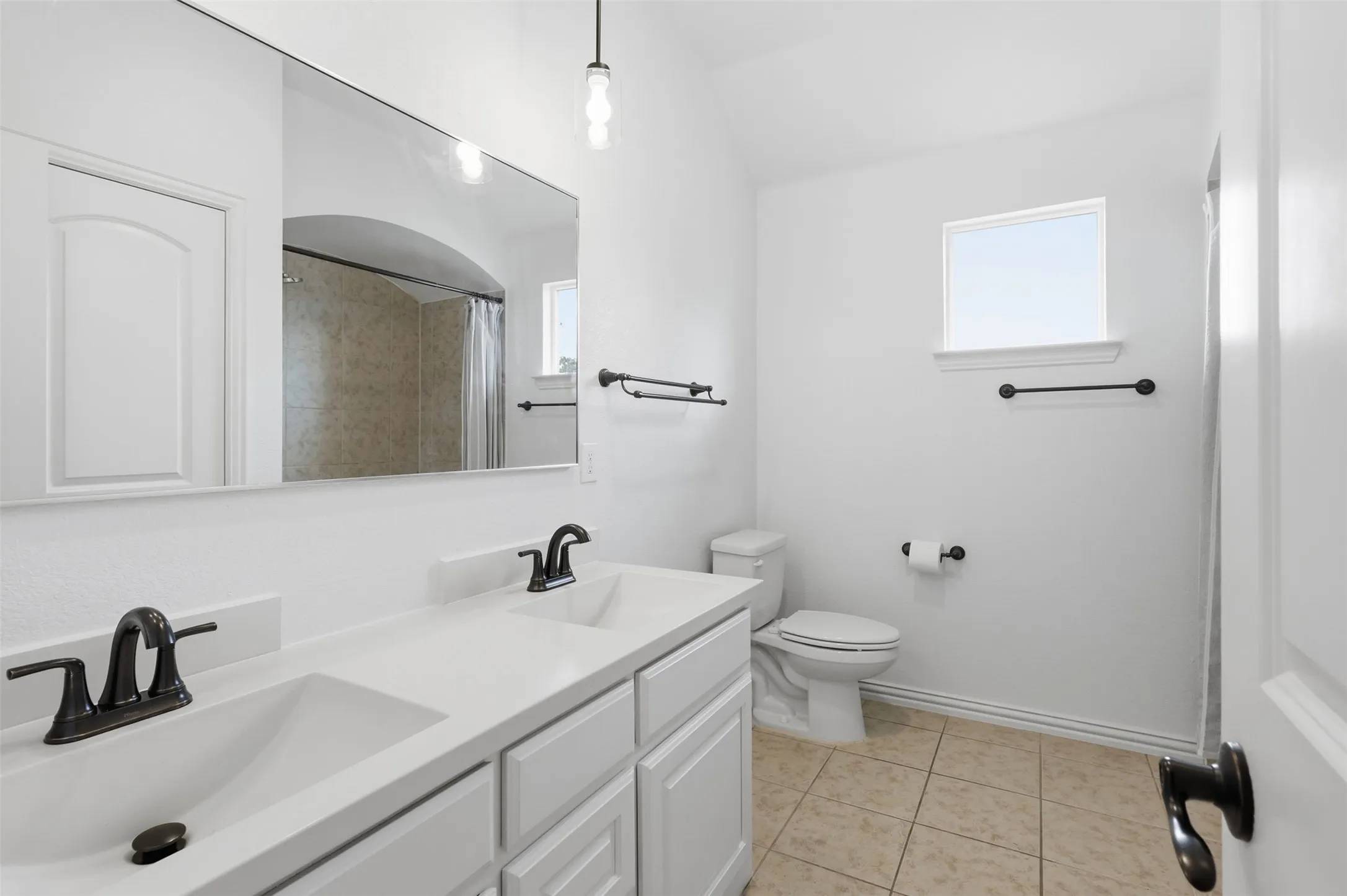 Dual-sink vanity with white cabinetry, light-toned solid surface countertops, oil-rubbed bronze fixtures, large vanity mirror, and ceramic tile flooring