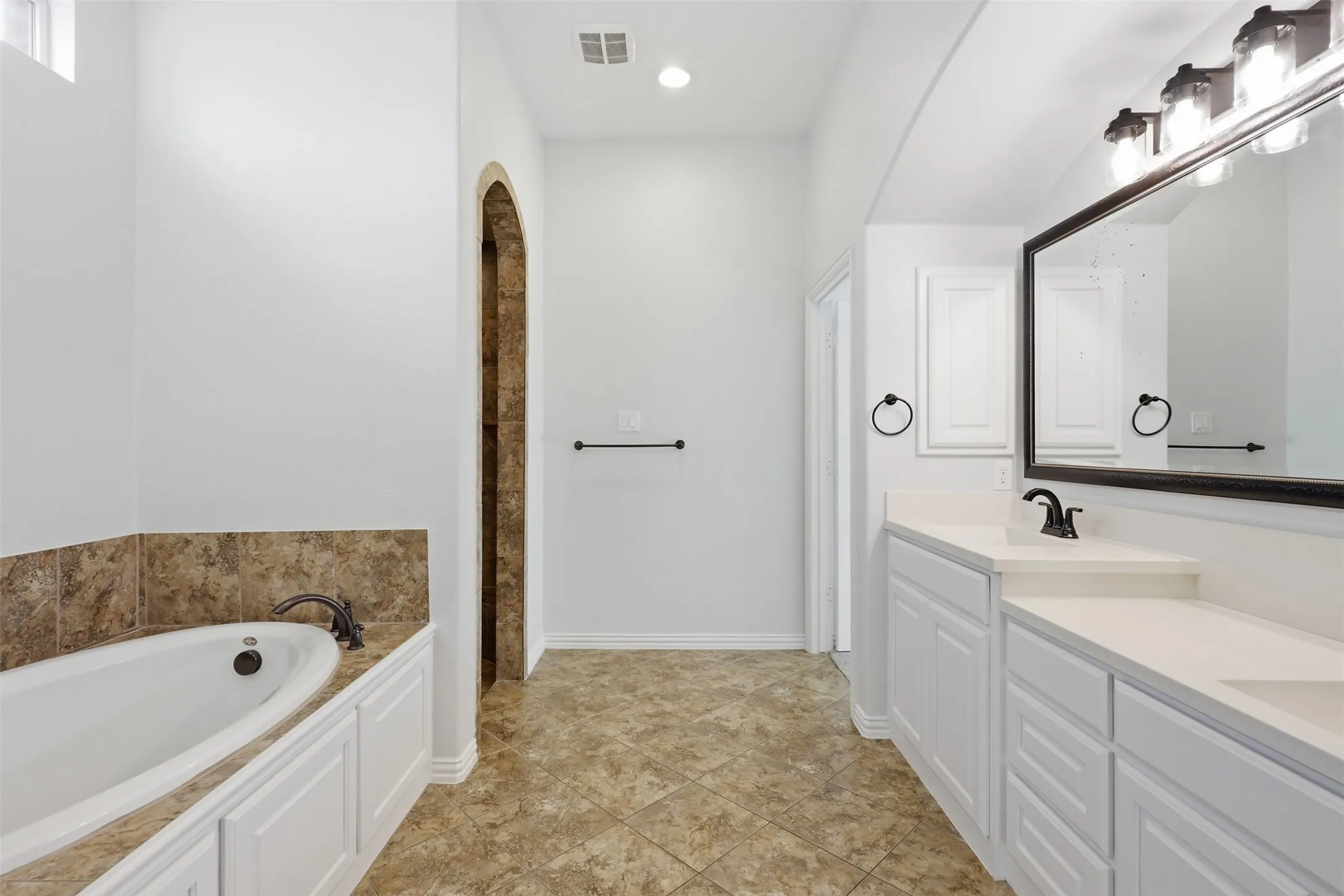 Bathroom featuring a dual-sink vanity with white cabinetry, quartz countertop, framed wall mirror, tiled bathtub surround, and arched tiled shower entry