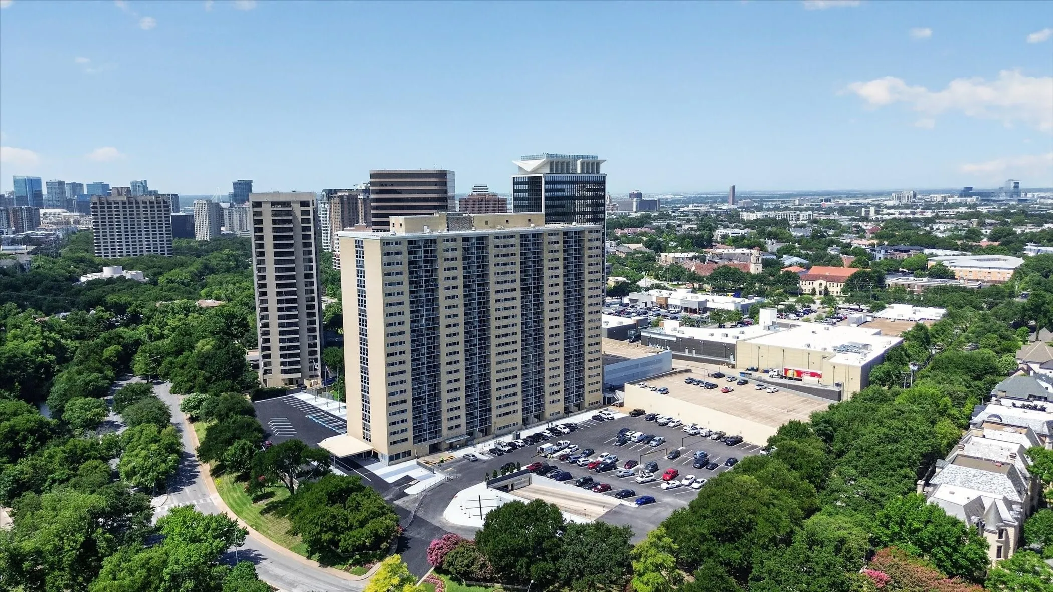 Aerial perspective of high-rise building with an asphalt parking lot, paved surface-level driving lanes, and mature surrounding tree canopy