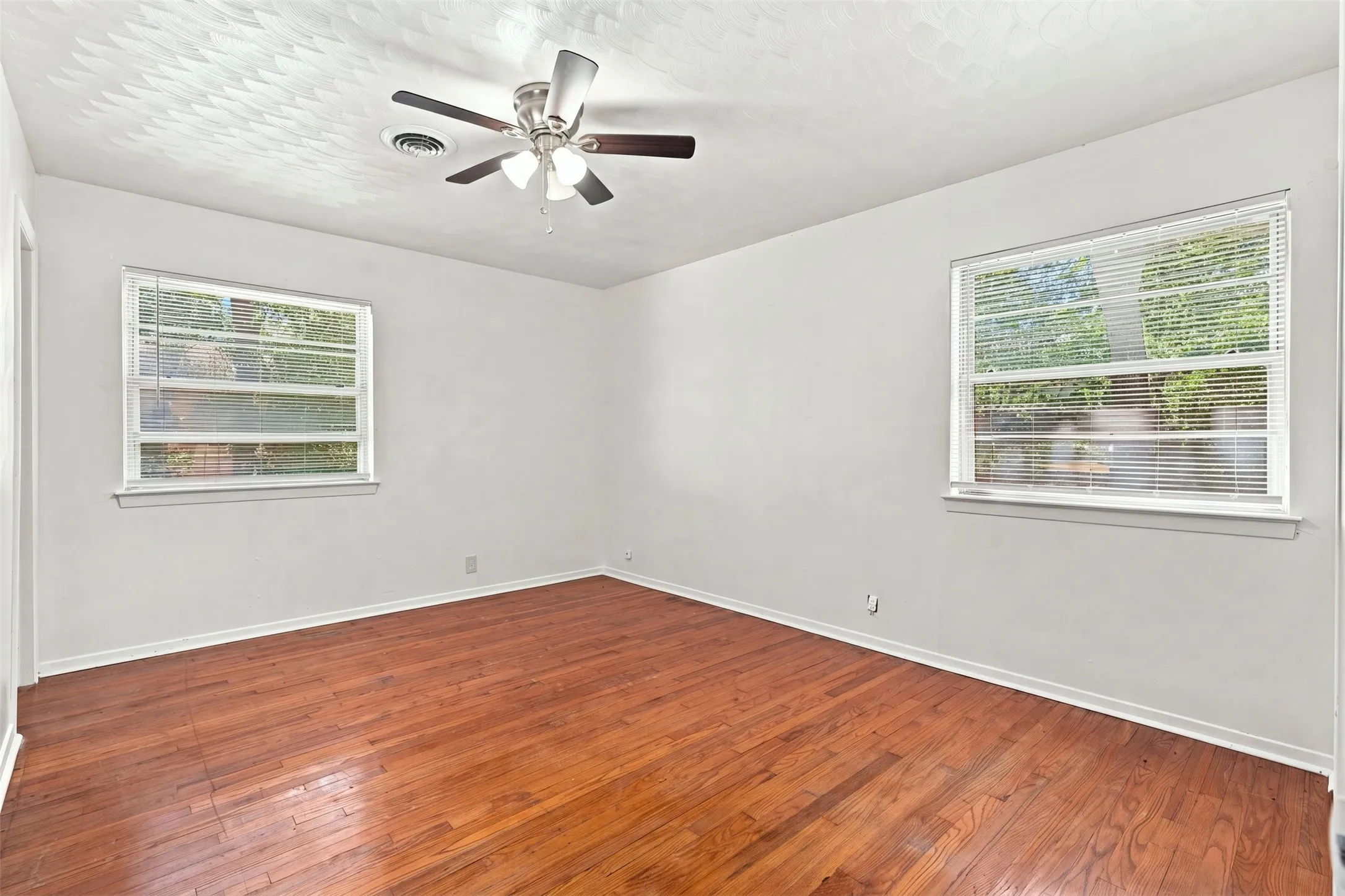 Room featuring rich wood-finish flooring, two windows with blinds, a ceiling fan with integrated lighting, a ceiling vent, and light-toned wall finishes