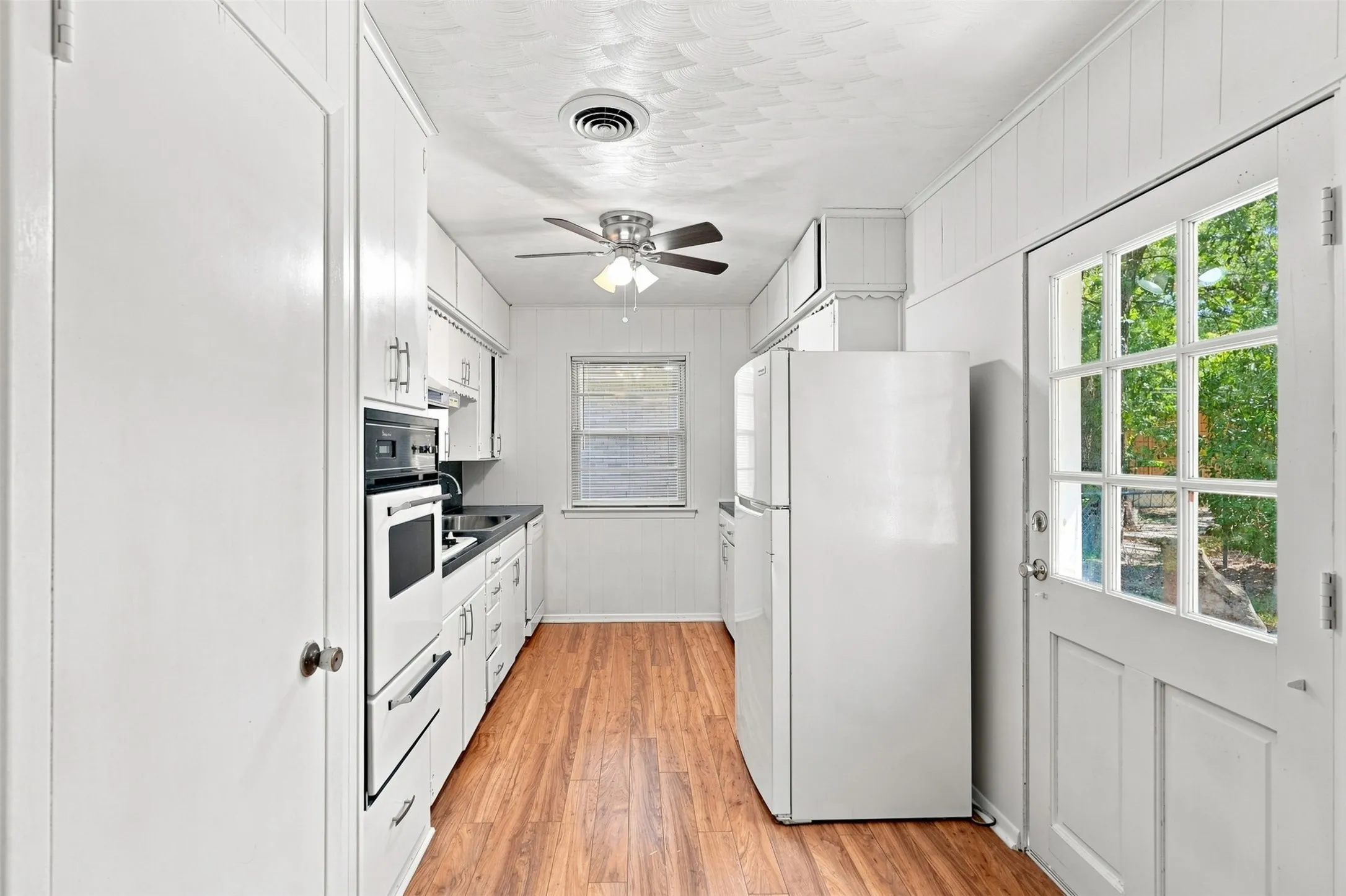 Galley kitchen featuring wood-finish flooring, white cabinetry with contemporary hardware, integrated wall oven, and a paneled glass-insert door