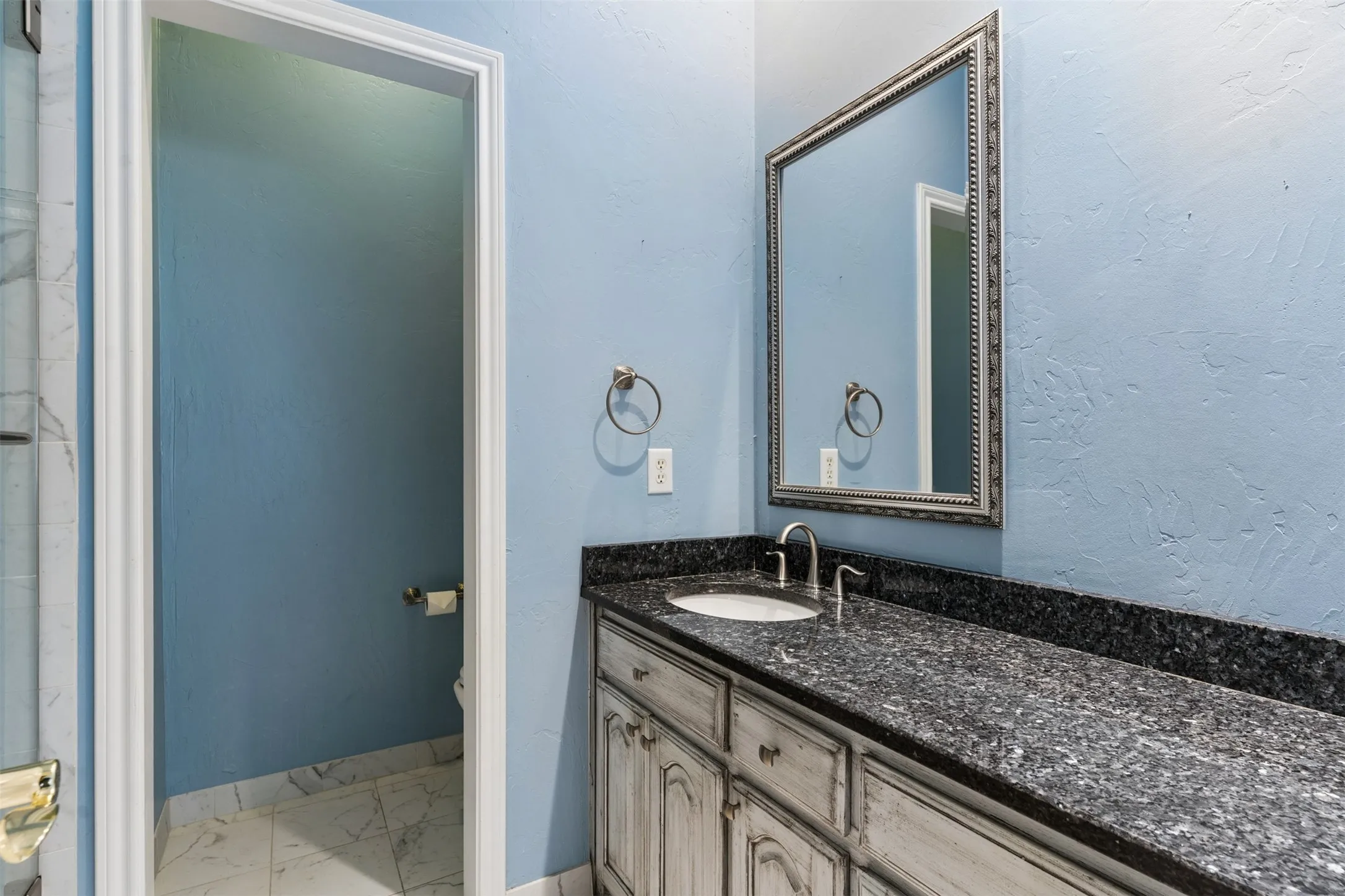 Custom cabinetry with a distressed finish, dark stone-top vanity, undermount sink, arched chrome faucet, and rectangular framed mirror