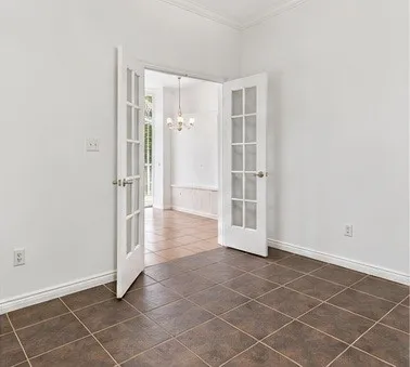 White-painted interior space featuring crown molding, ceramic tile flooring, and glass-paneled French doors leading to a secondary room with a built-in cabinet and a hanging light fixture