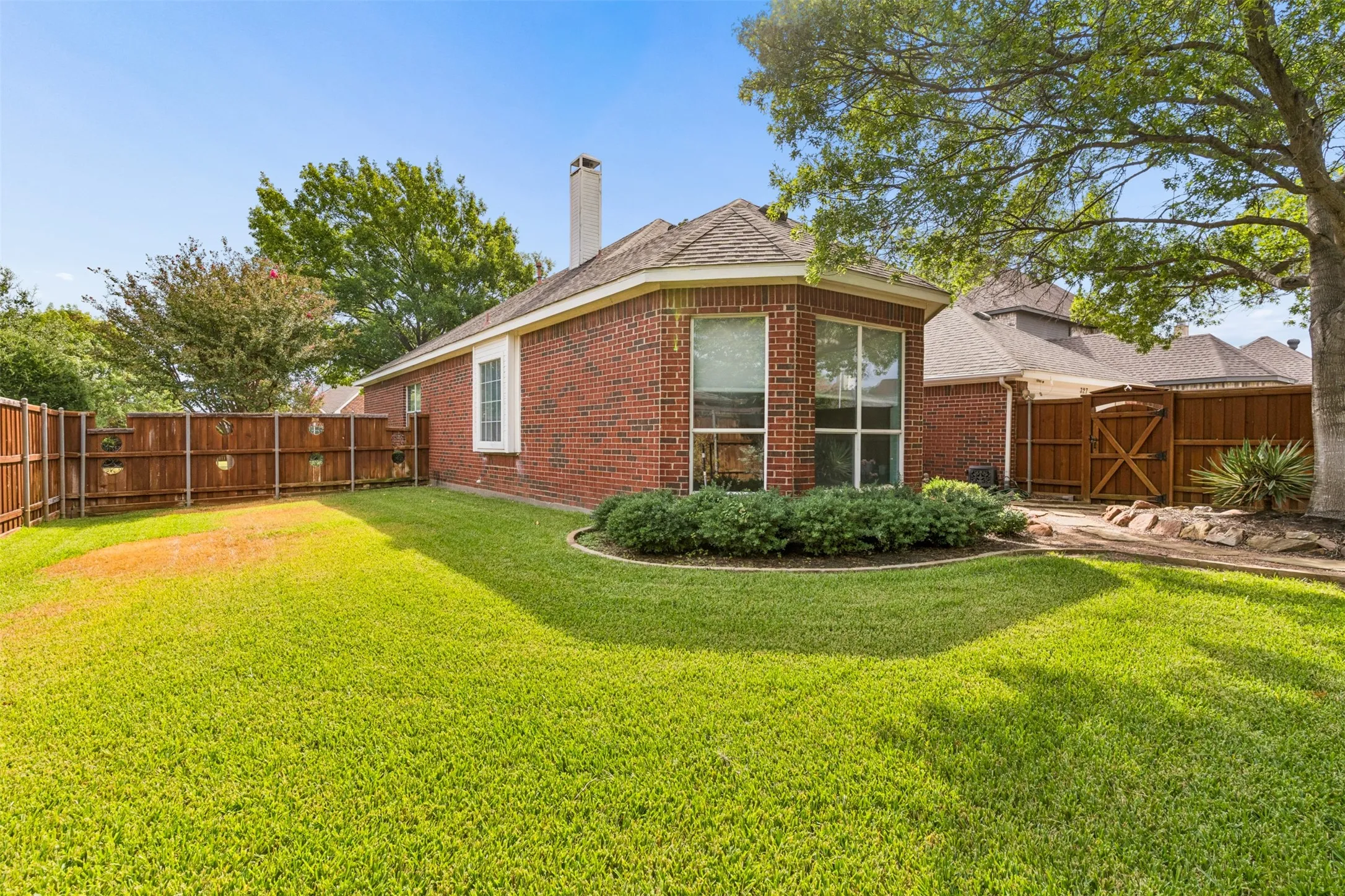 Brick exterior featuring a multi-faceted roofline, chimney, expansive lawn, mature trees, and wooden perimeter fencing