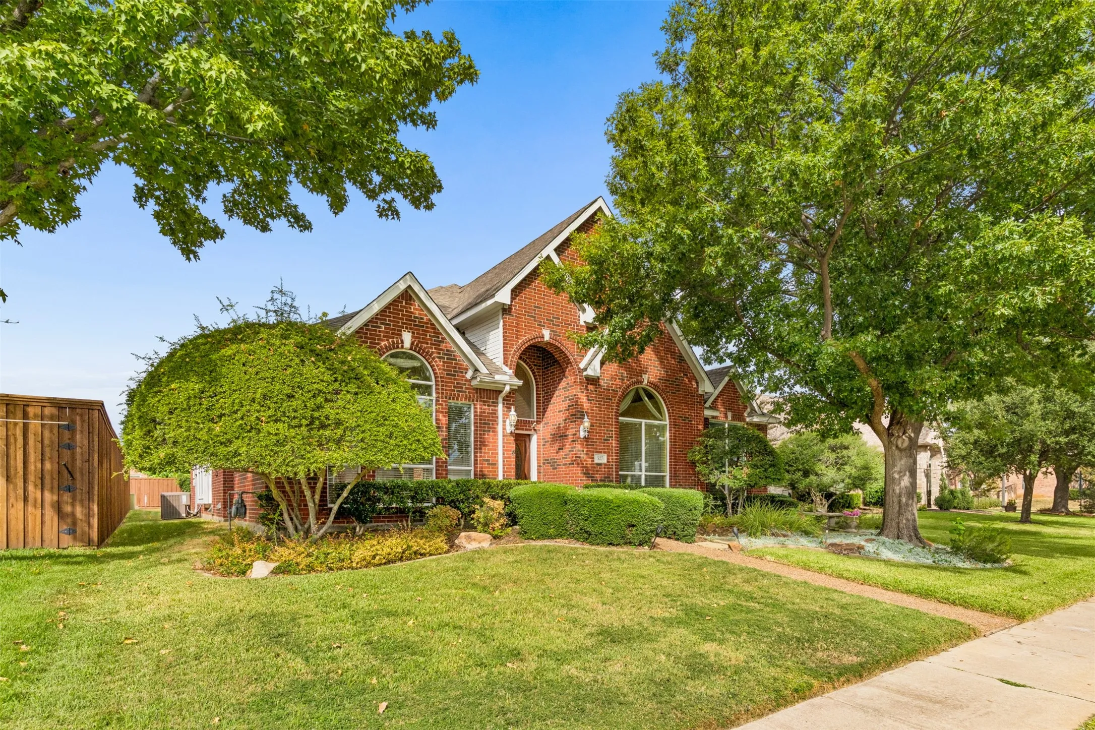 Brick exterior featuring an arched entryway, steep roof gables, and large arched windows, set on a landscaped lot with mature trees