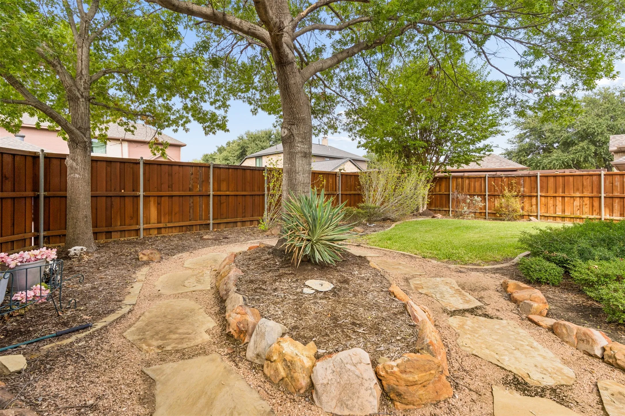 Backyard featuring a wood privacy fence, stone paver pathway, elevated stone-bordered garden bed, and mature trees