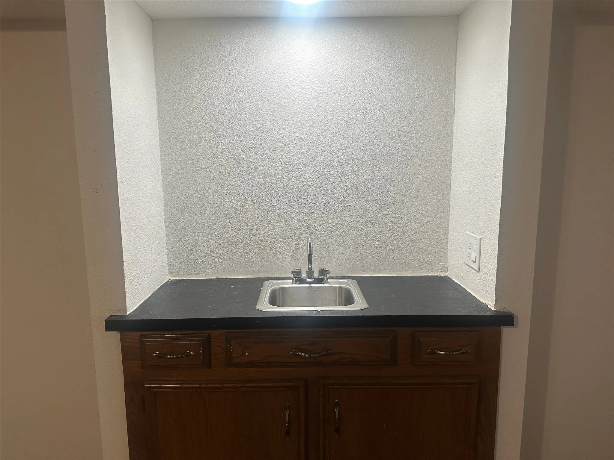 Built-in wet bar featuring a recessed alcove, dark countertop, stainless steel drop-in sink, and wood-finish cabinetry