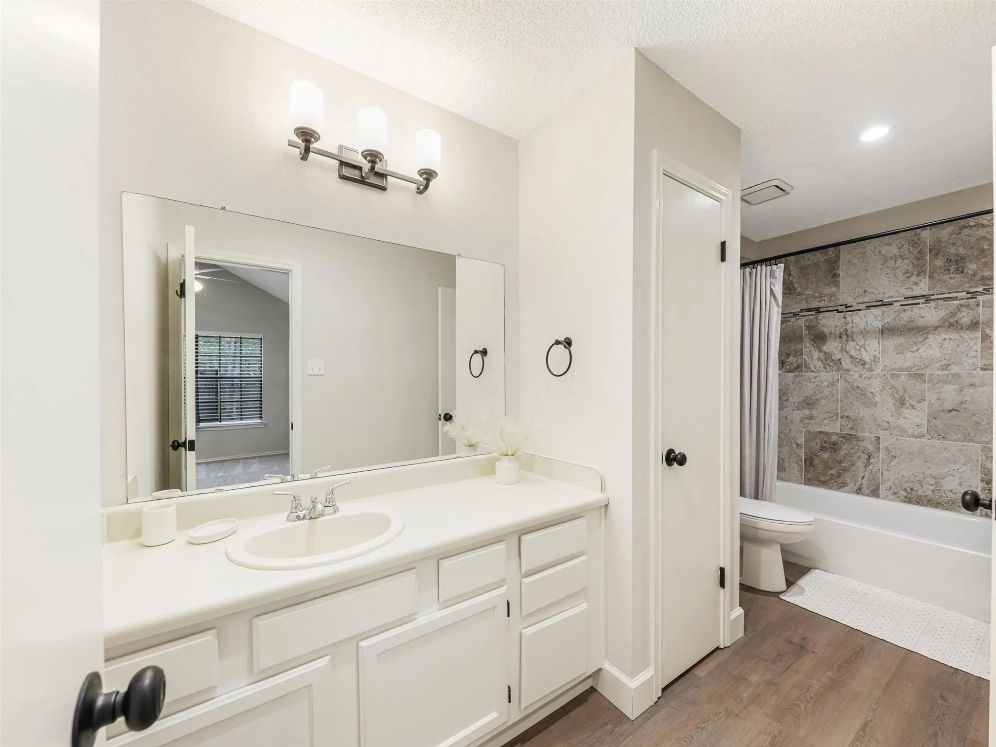 White vanity featuring a molded sink and integrated countertop, full-width wall mirror with three-light fixture, wood-finish flooring, and bathtub surround accented with large-format stone-look tile