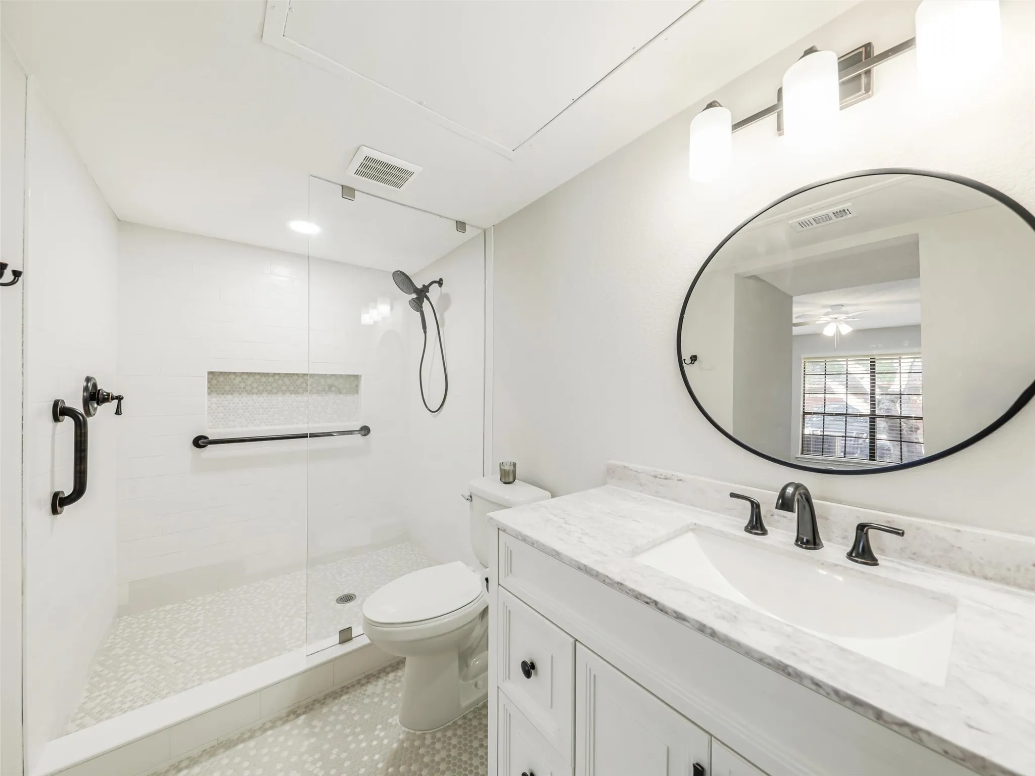 White vanity with a stone countertop, oval mirror, walk-in shower with a glass panel, stone tile flooring, and matte black fixtures
