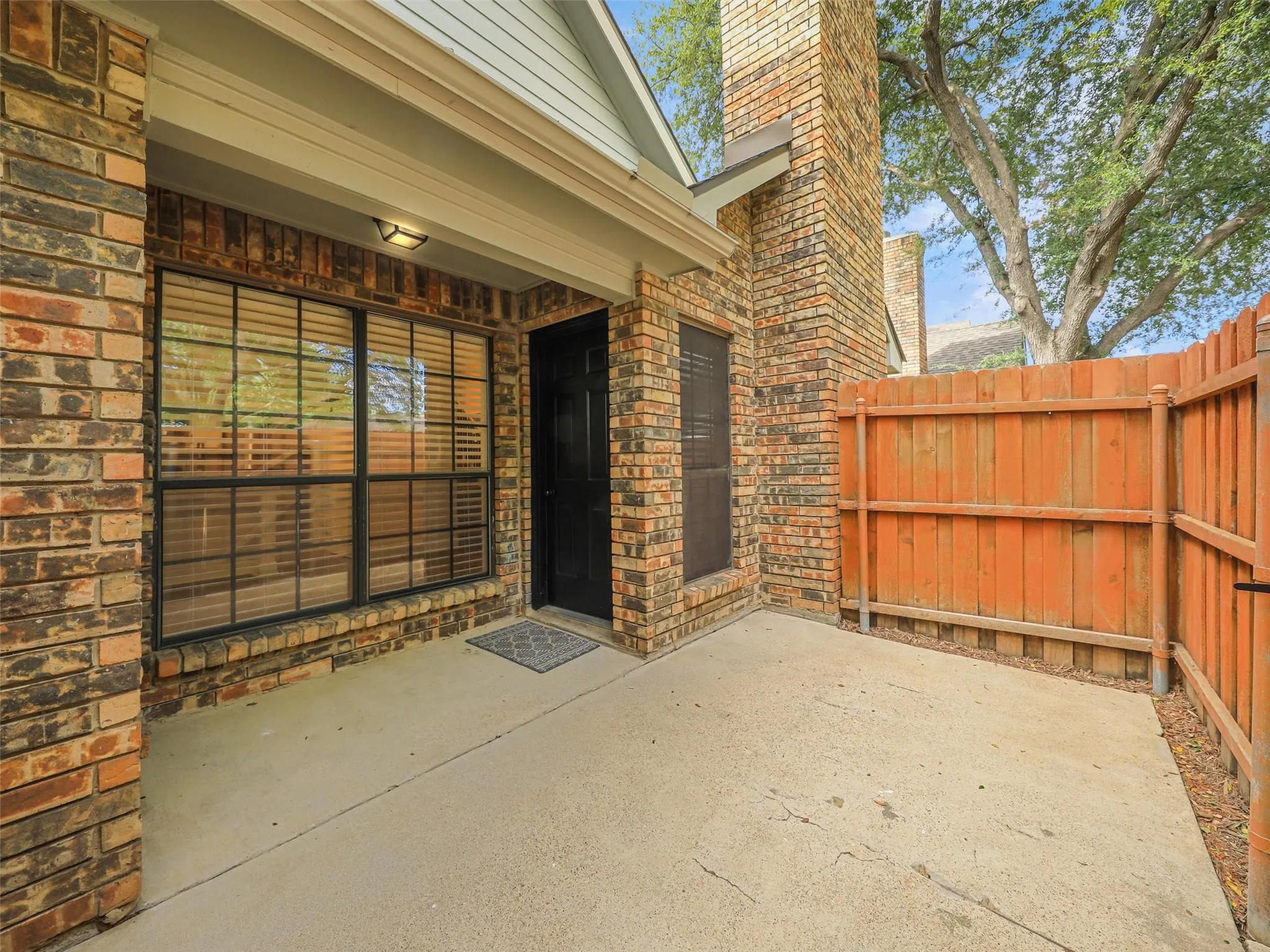 Brick exterior patio featuring a concrete slab, floor-to-ceiling brick chimney, dark-trimmed windows, and a wood privacy fence