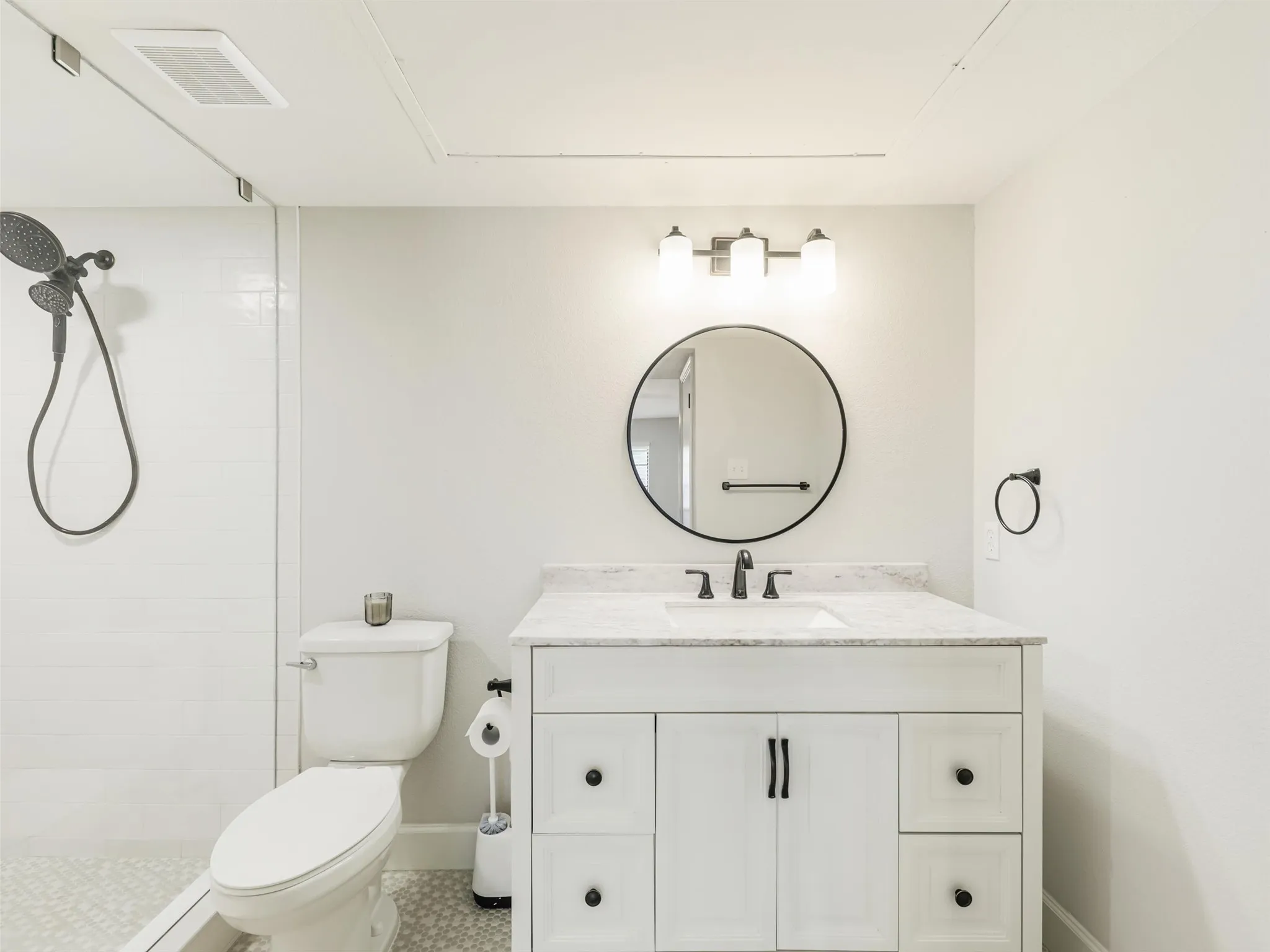 Bathroom featuring a white vanity with stone-top surface, undermount sink, round wall-mounted mirror, three-light fixture, and a glass-enclosed shower with handheld sprayer