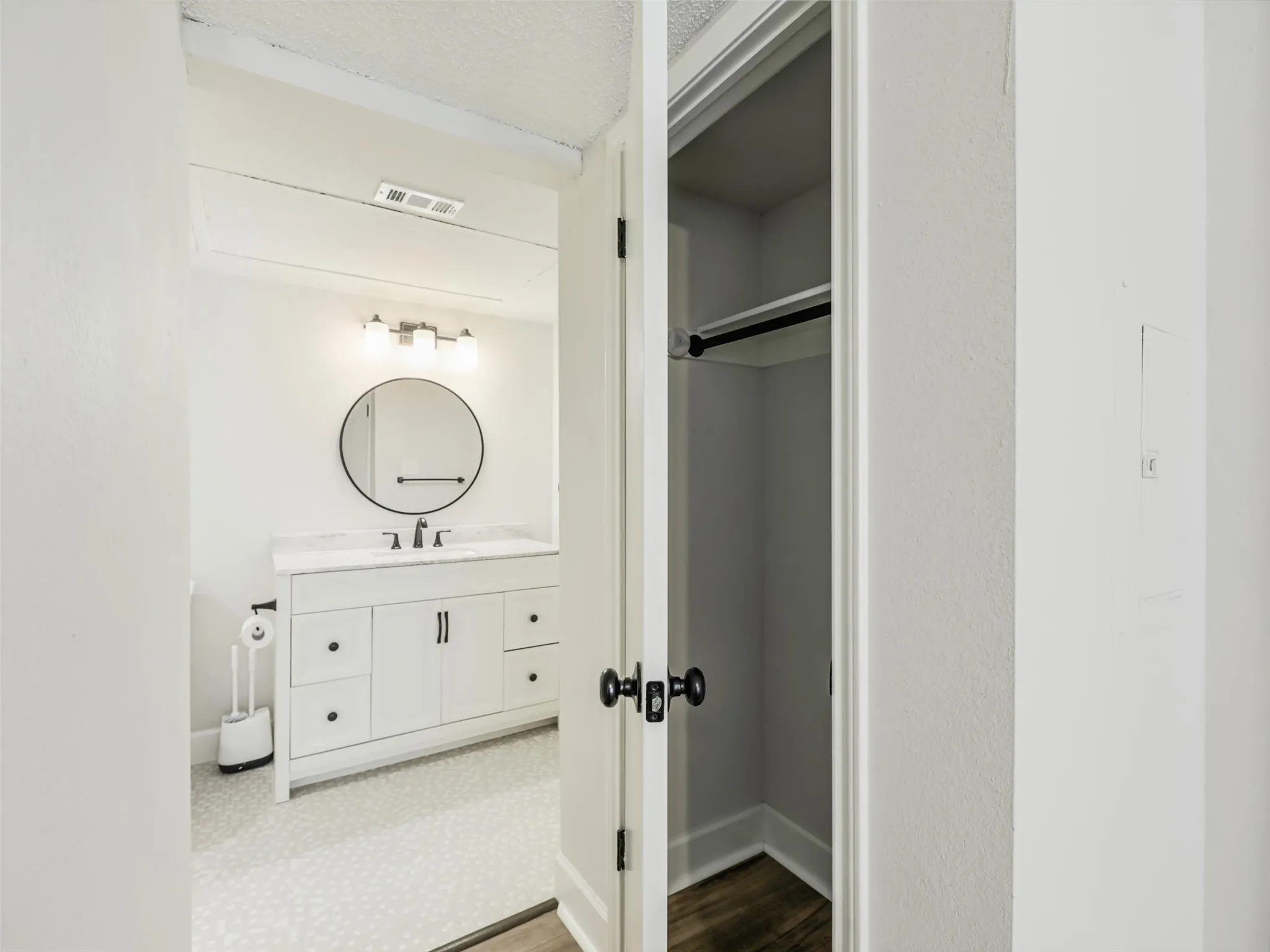 Bathroom featuring a white vanity with drawers, a round mirror, three-light fixture, and patterned flooring, complemented by a storage closet with a built-in rod and shelf