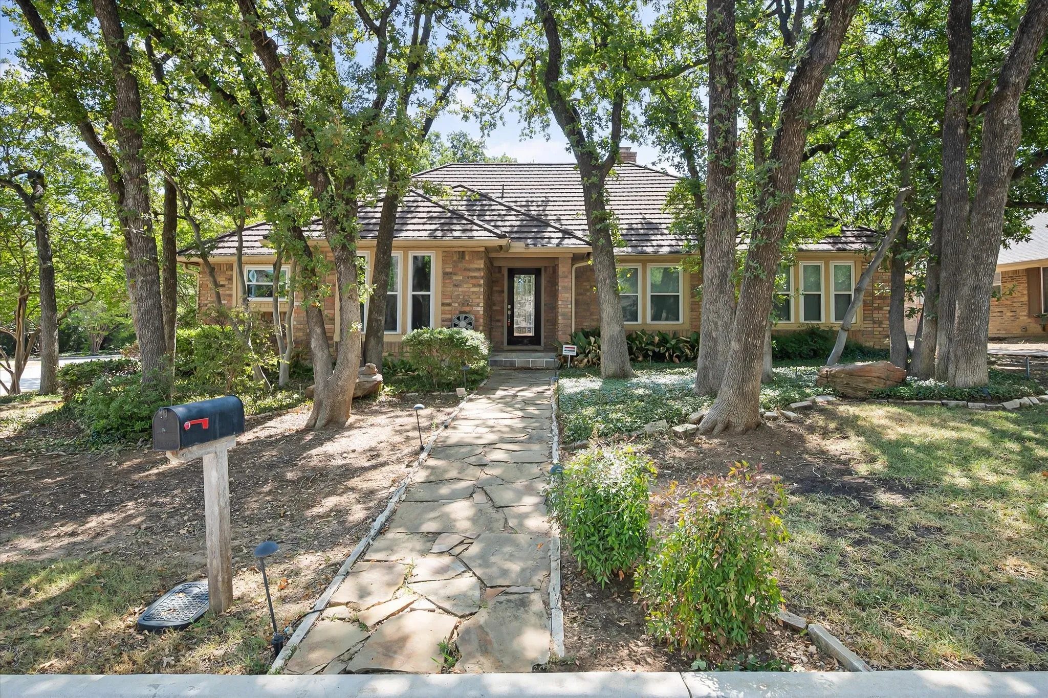 Brick exterior residence featuring a flagstone walkway, metal roofing, large mature trees, and landscaping