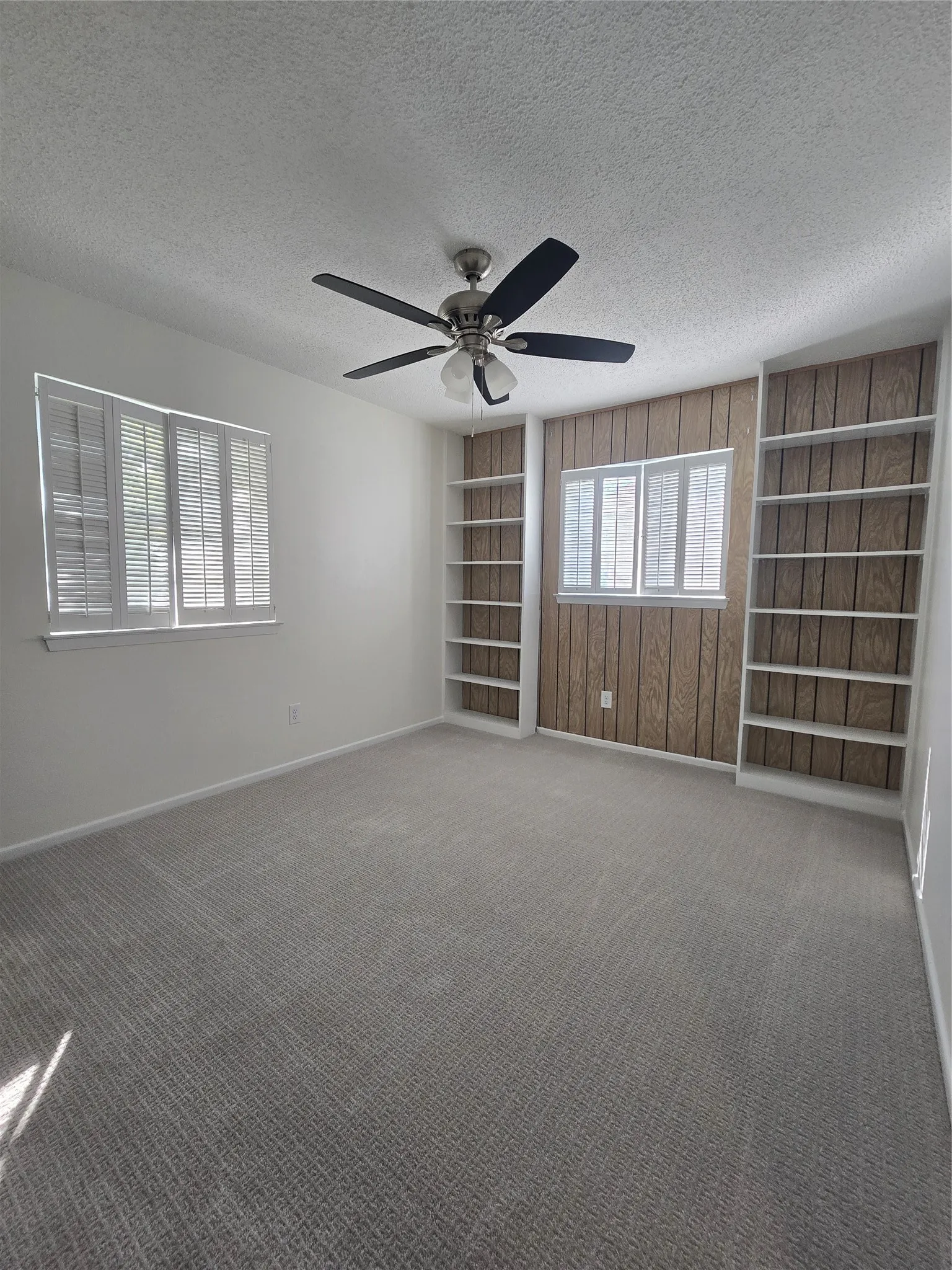 Room featuring wall-to-wall carpeting, ceiling fan, wood-paneled accent wall with built-in shelving, and white plantation-style shutters