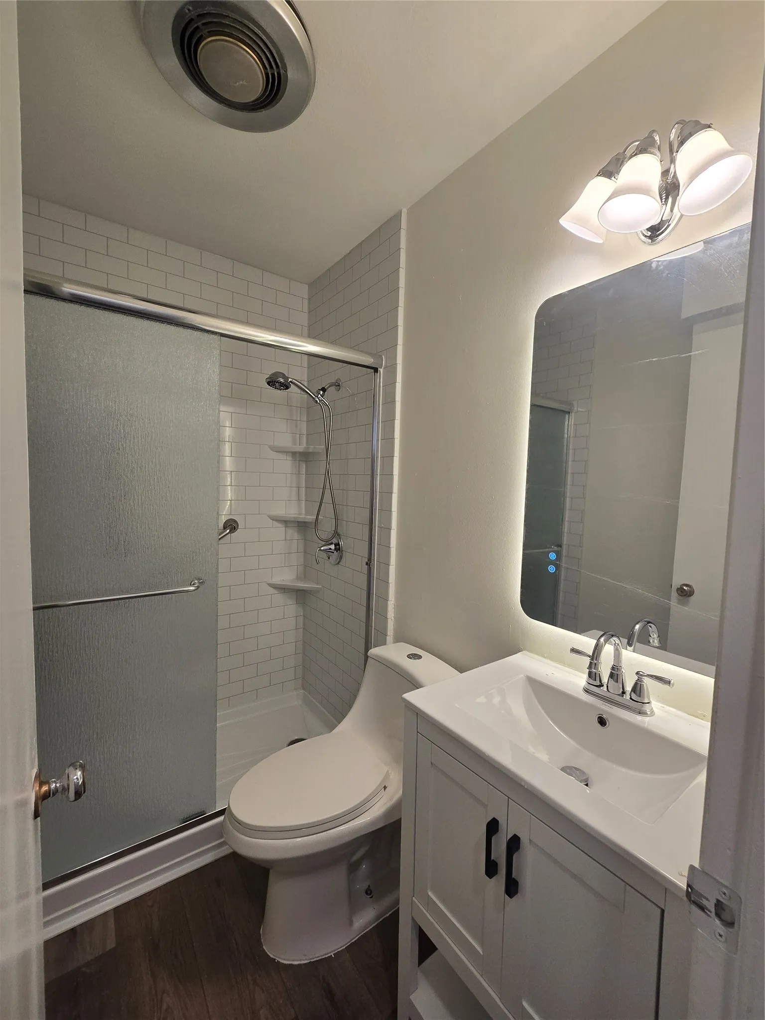 Bathroom featuring a white vanity with an integrated sink, a rectangular mirror with integrated perimeter lighting, a walk-in shower with white subway tile surround, and wood-finish flooring