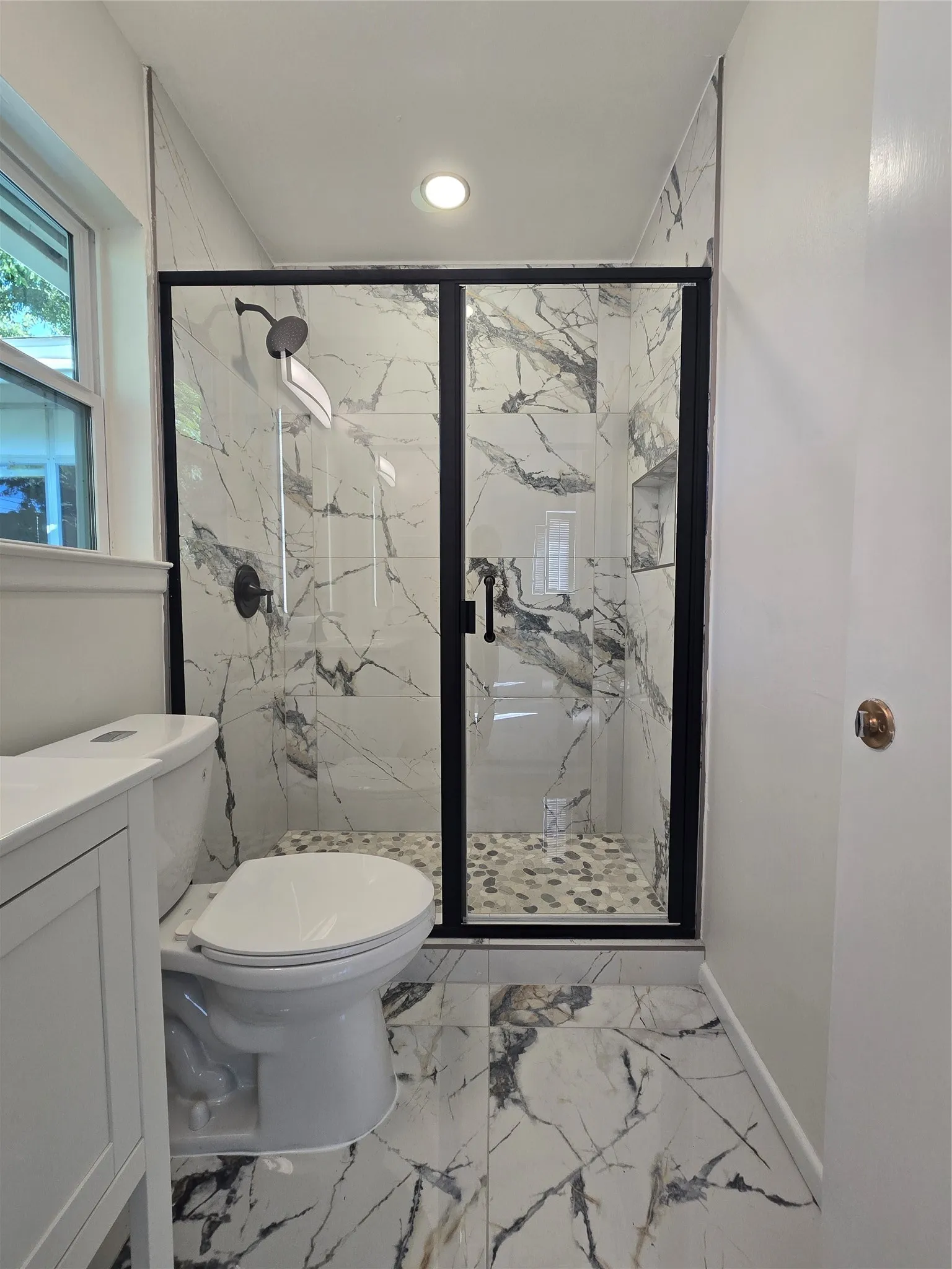 Walk-in shower featuring large-format patterned wall tile, black metal frame, integrated recessed niche, and pebble-tile flooring, complemented by a white vanity and matching large-format floor tile