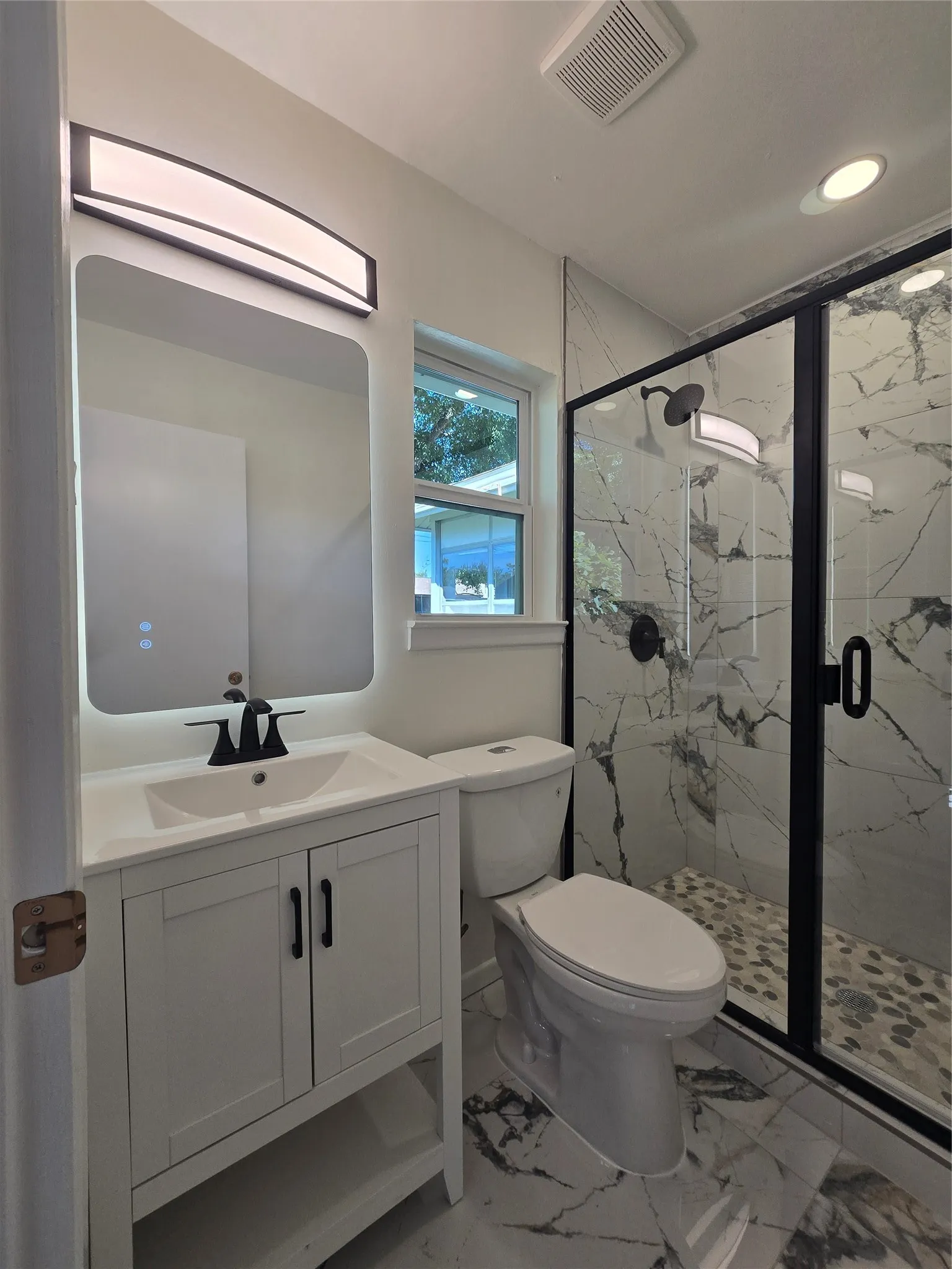 White vanity cabinet with a vessel sink and matte black hardware, backlit rectangular mirror, and a glass-enclosed shower featuring stone-patterned wall tile and pebble flooring
