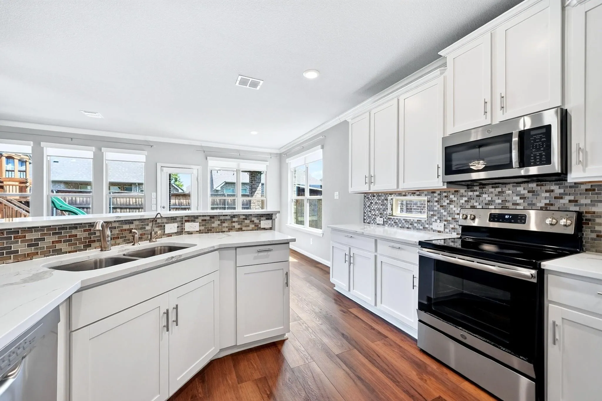 Kitchen featuring white shaker-style cabinetry, stainless steel appliances, multi-tonal glass tile backsplash, light-colored countertops, and wood-finish flooring