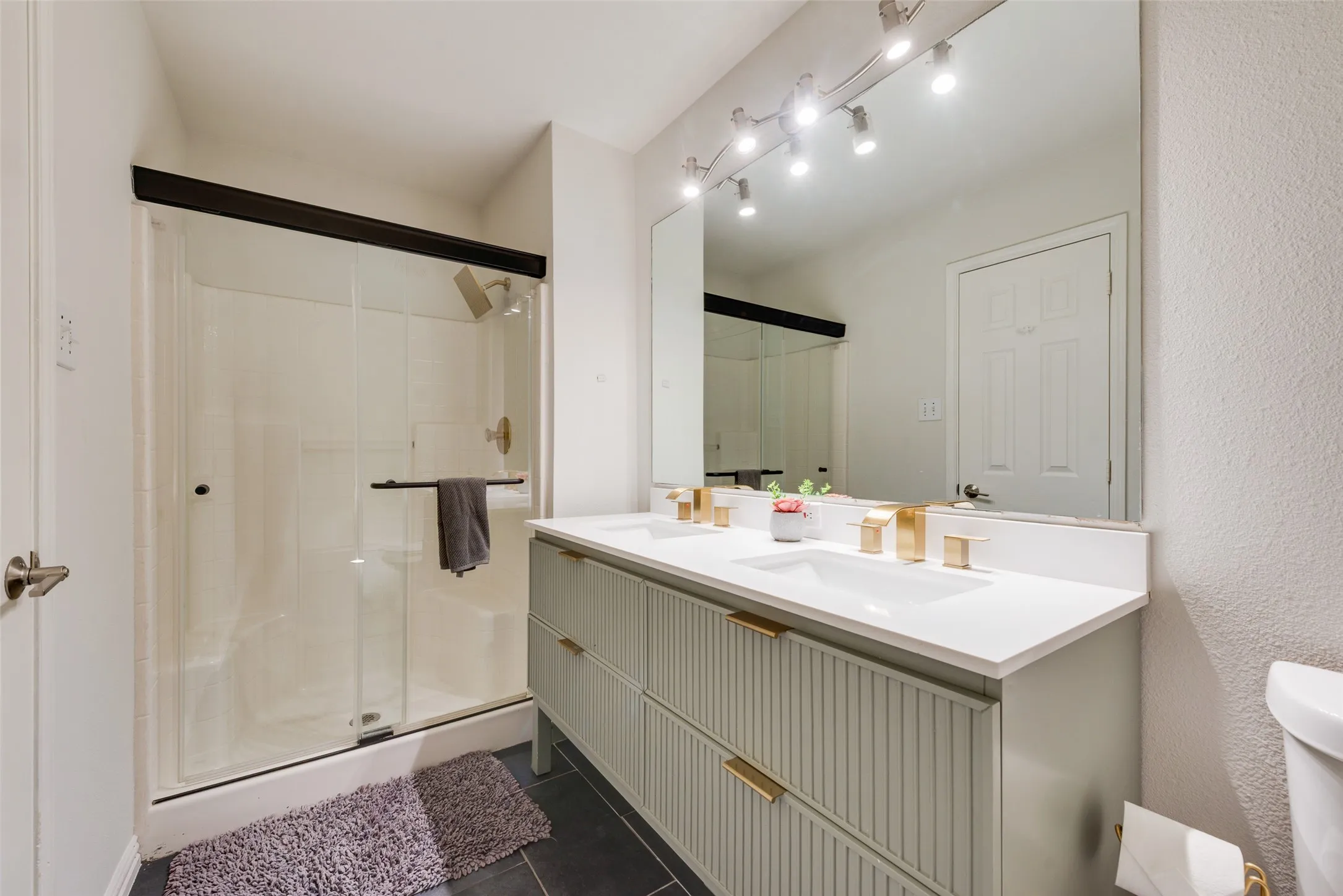 Double vanity featuring a fluted-style cabinet base, stone countertop, integrated rectangular sinks, and metallic finish hardware, complemented by a walk-in shower with a glass enclosure