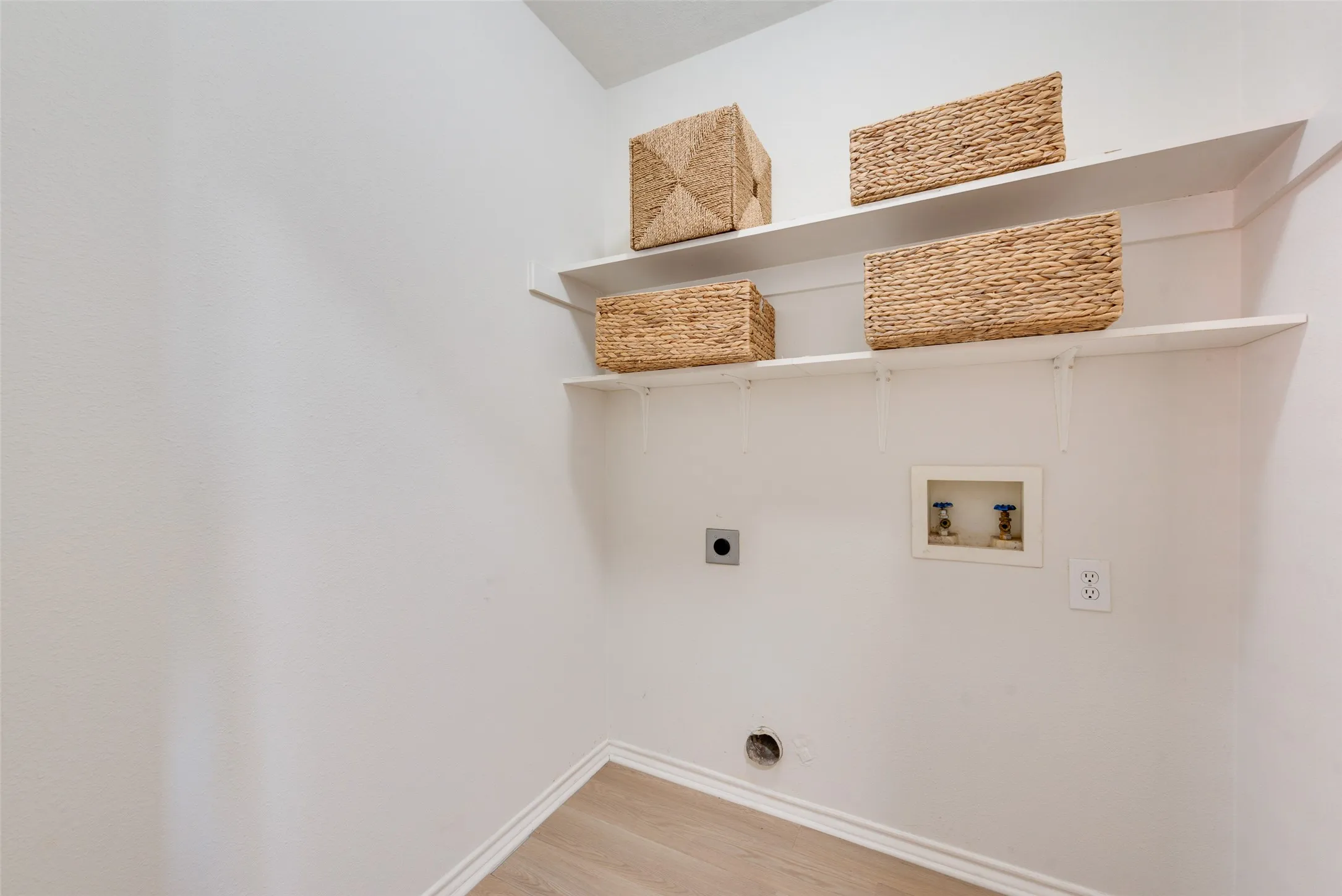 Utility space featuring two tiered wall-mounted shelves, wood-finish flooring, and dedicated water and electrical hookups