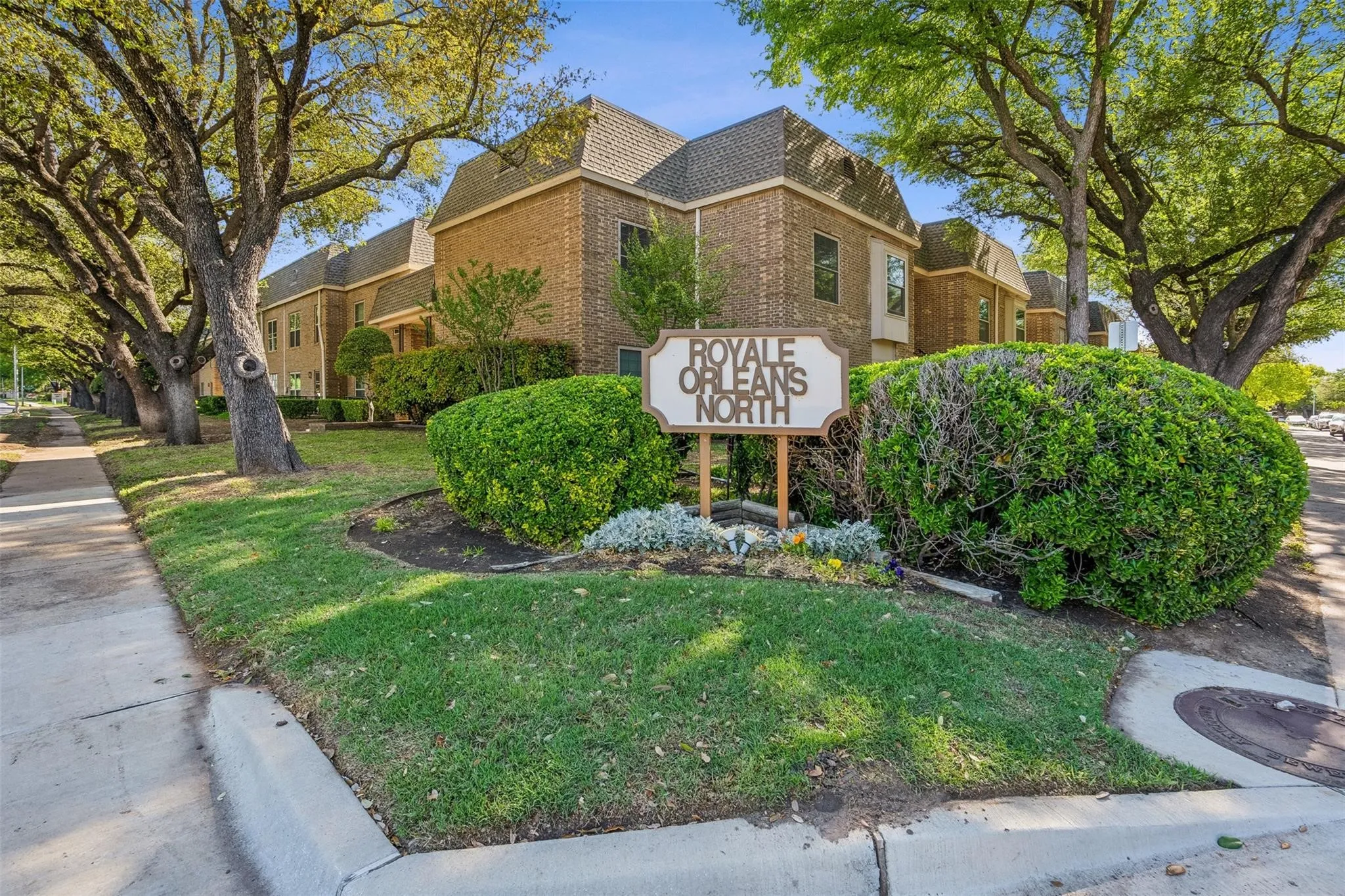 Multi-story brick exterior featuring a hip roof, mature trees, manicured shrubbery, and concrete sidewalks