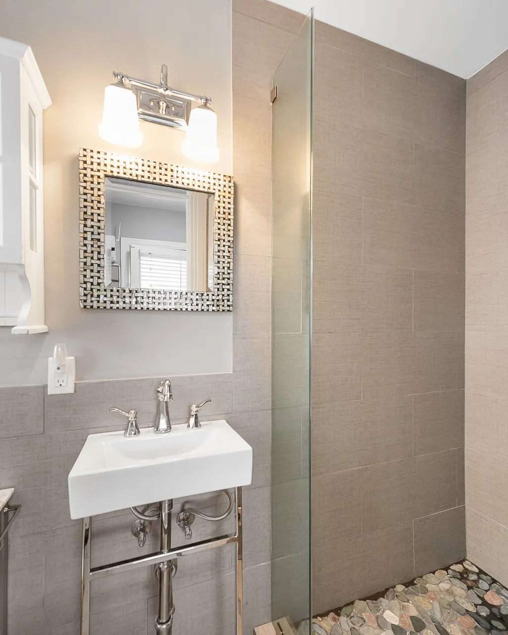 Wall-mounted console sink with chrome finish, textured tile wall surround, frameless glass shower panel, pebble stone flooring, and polished chrome light fixture
