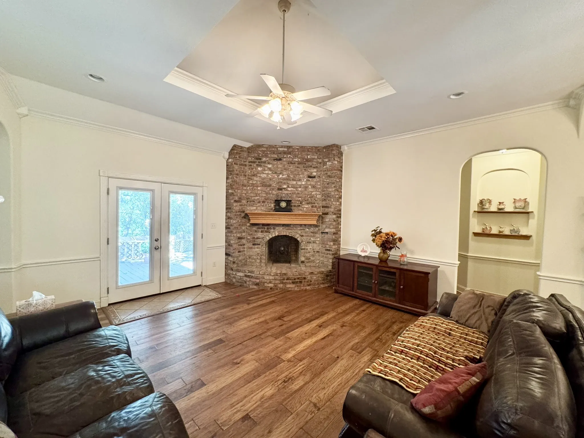 Vaulted ceiling with crown molding, corner brick fireplace featuring a wooden mantel, wood-finish flooring, double glass panel exterior doors, and a built-in arched wall niche with shelving