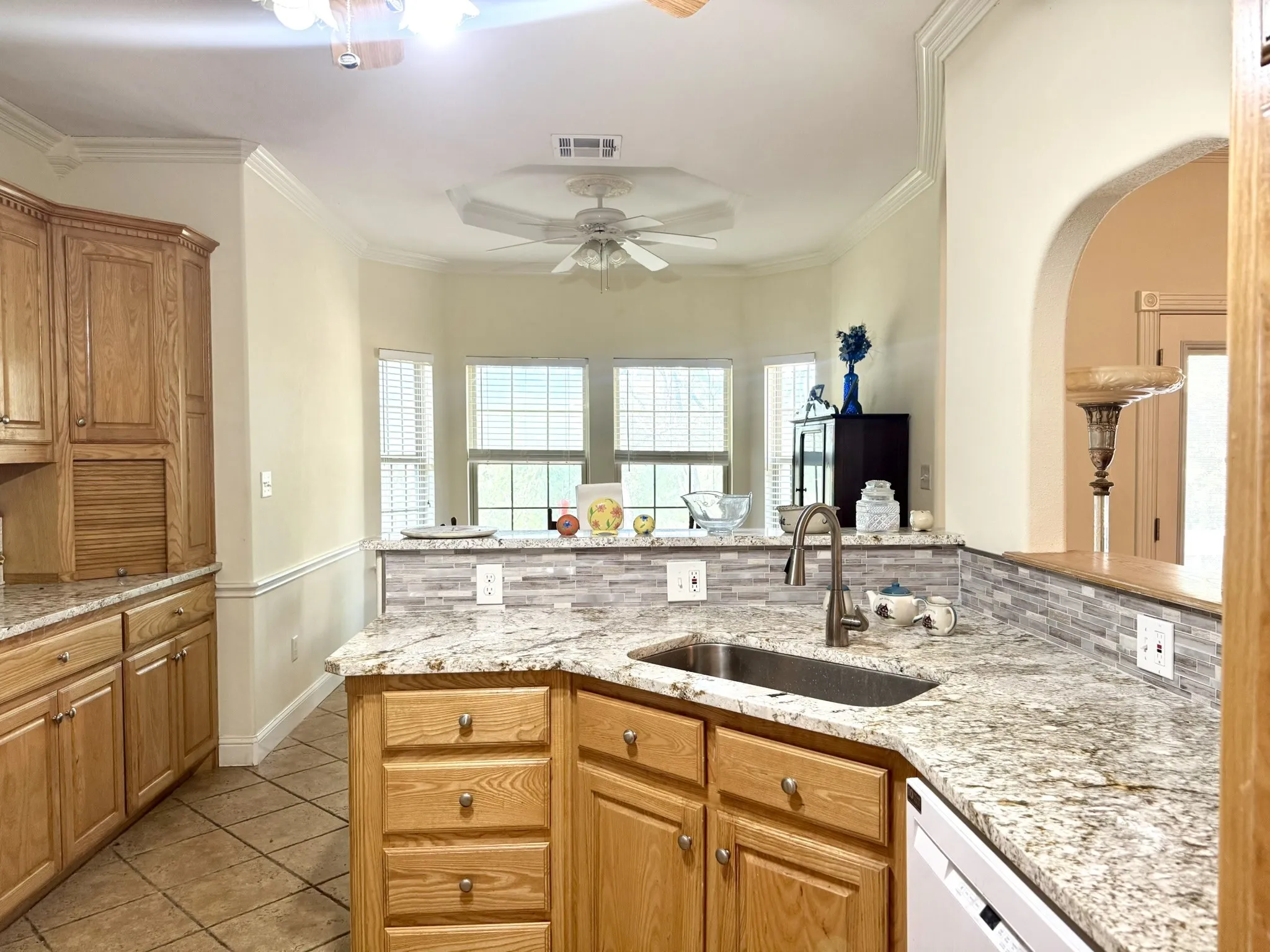 Kitchen featuring stone countertops, tile backsplash, natural finish wood cabinetry, under-mount sink with high-arch faucet, and crown molding