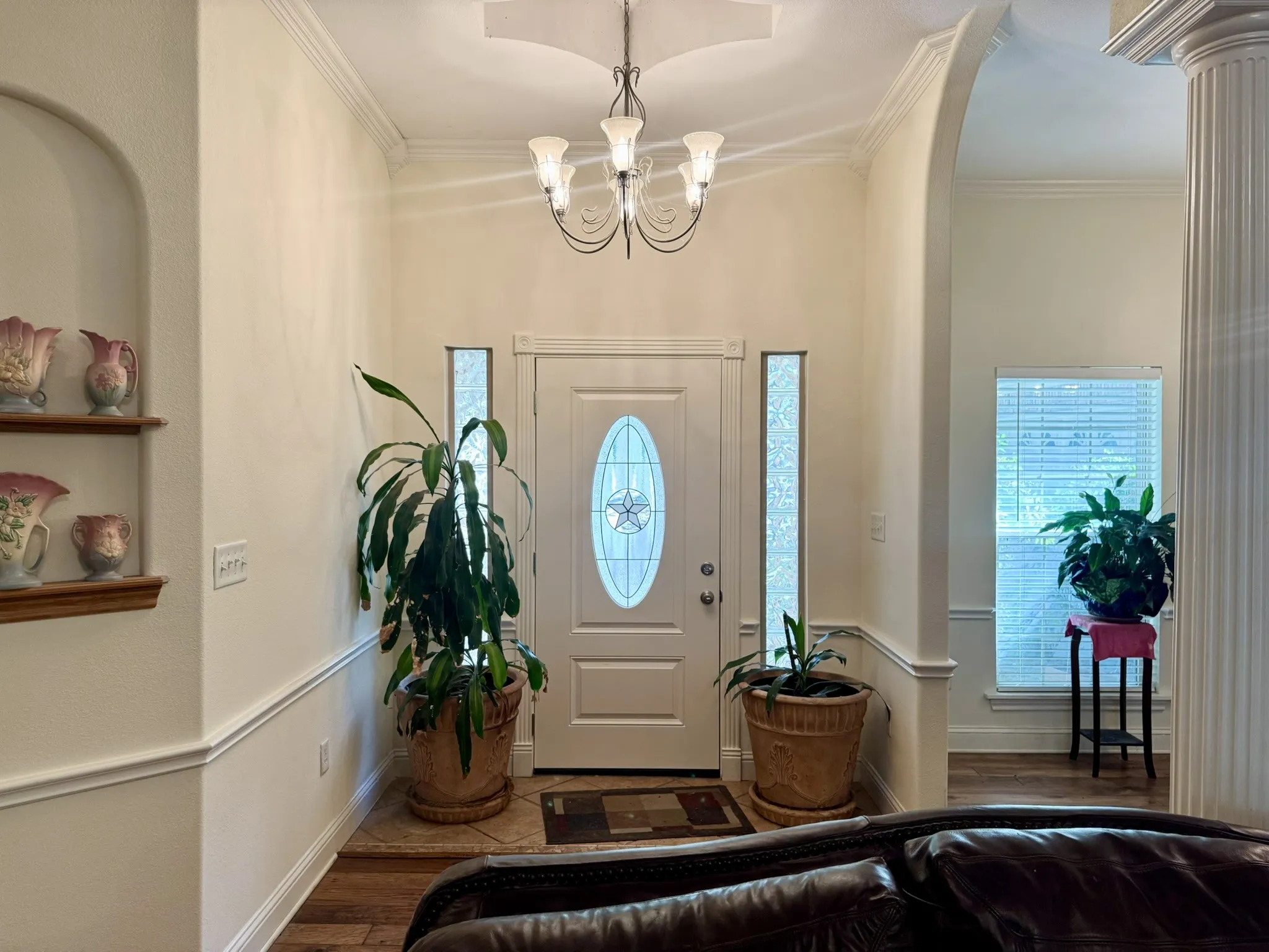 Entryway featuring arched architectural detailing, a central chandelier, decorative wall niche with built-in shelving, fluted column, and white door with oval glass insert flanked by narrow sidelights