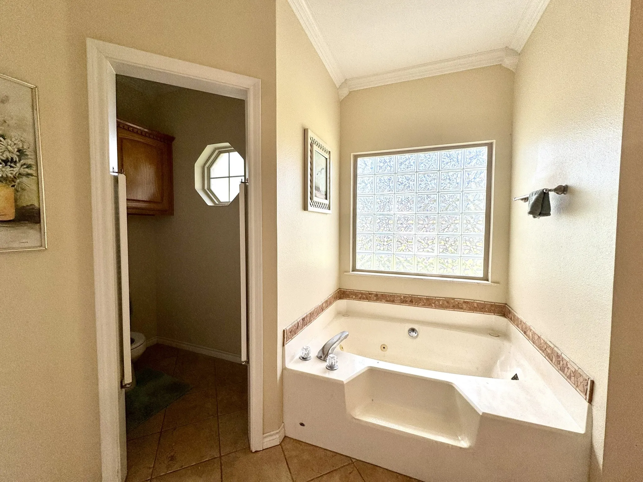 Corner jetted tub with tile surround, glass block window, crown molding, and tiled flooring