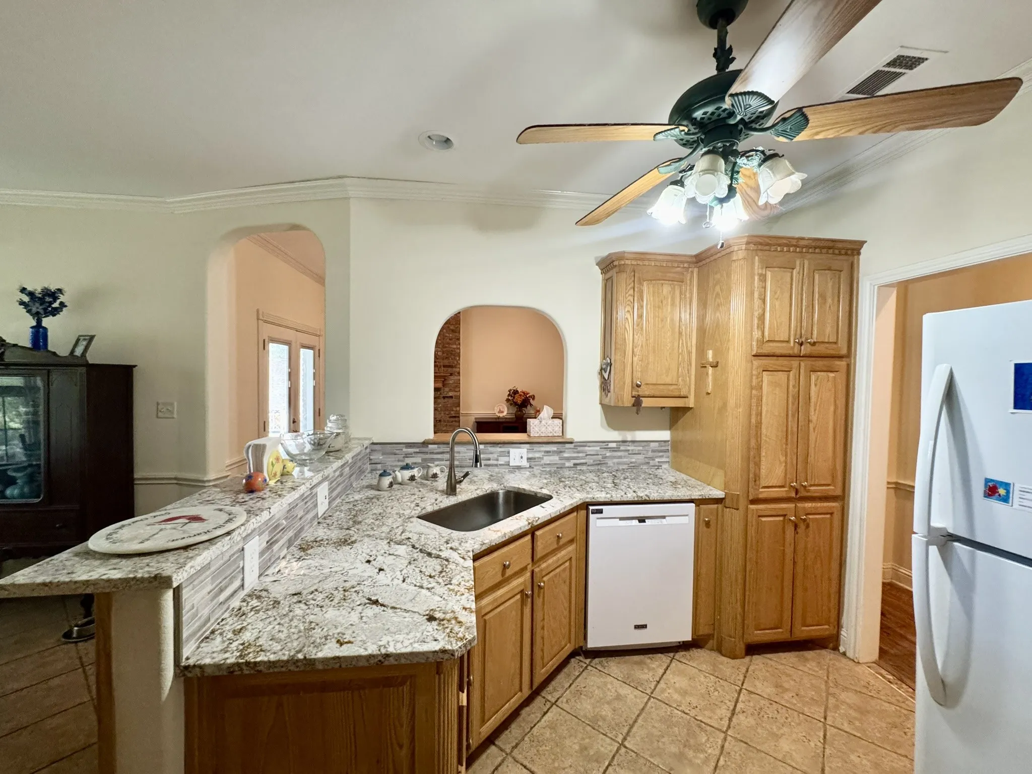 Kitchen featuring stone-finish countertops, tile backsplash, built-in light-tone wood cabinetry, tile flooring, and a ceiling-mounted fan with integrated lighting