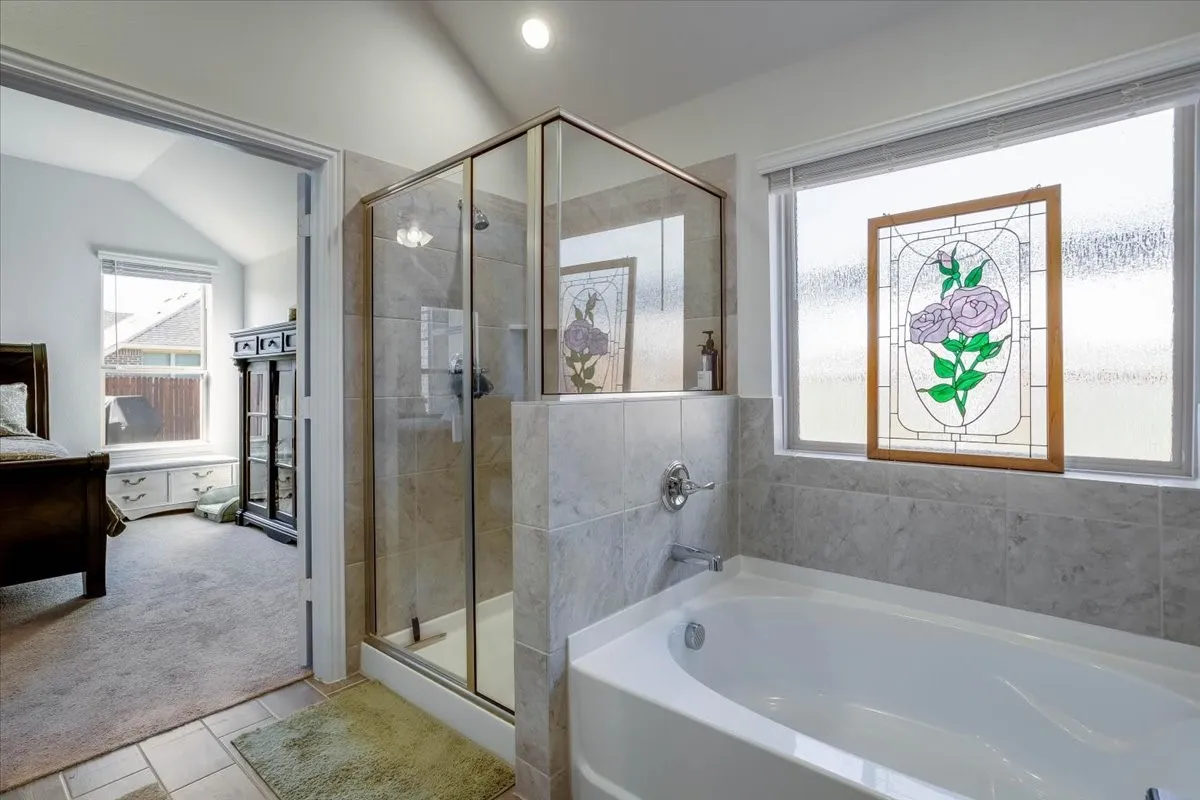 Frameless glass shower enclosure with neutral-toned tiled surround, deep soaking tub, polished chrome fixtures, and windows with stained glass decorative inserts
