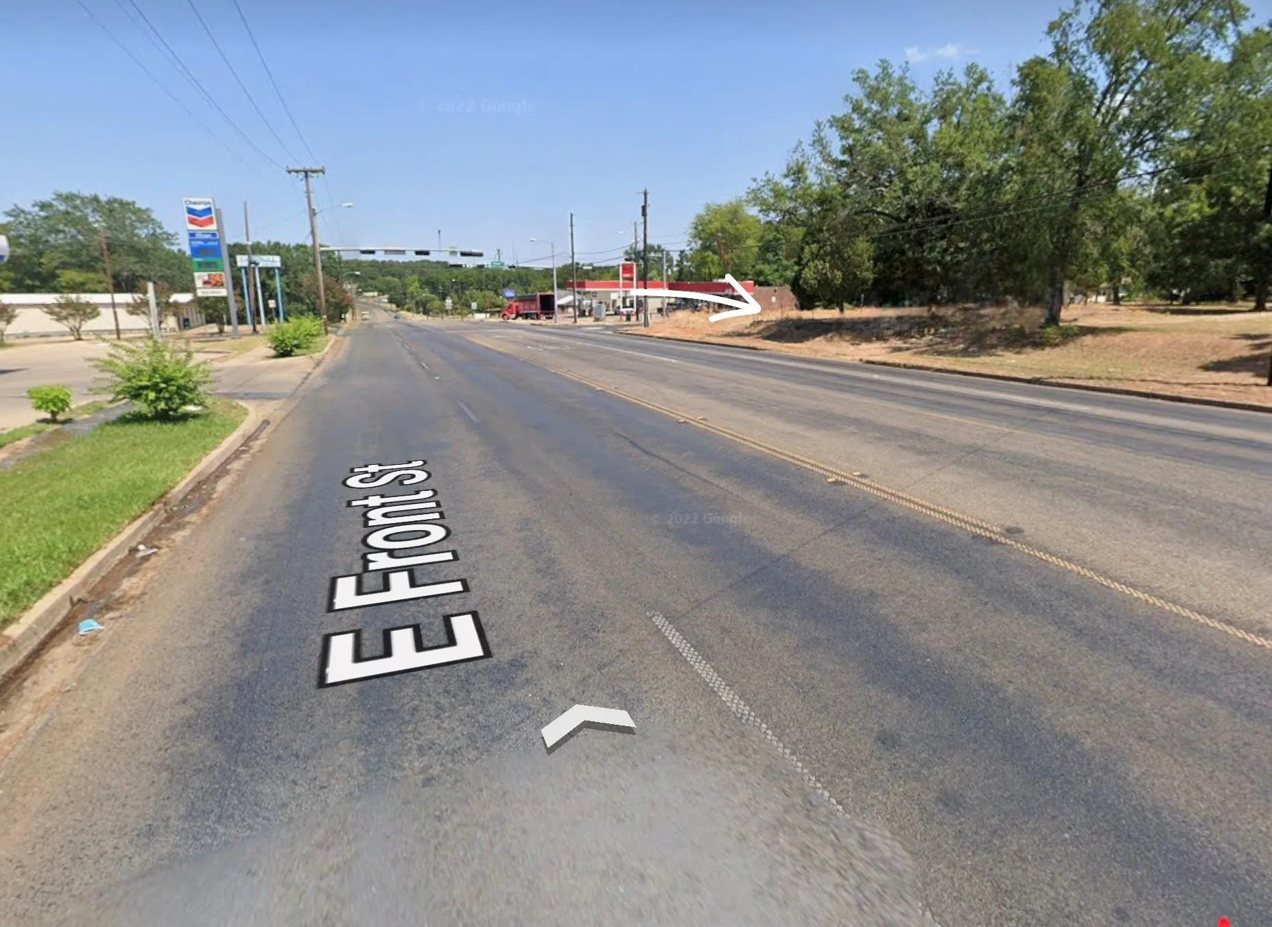 Street-level perspective featuring paved roadway frontage, cleared open lot, and mature trees